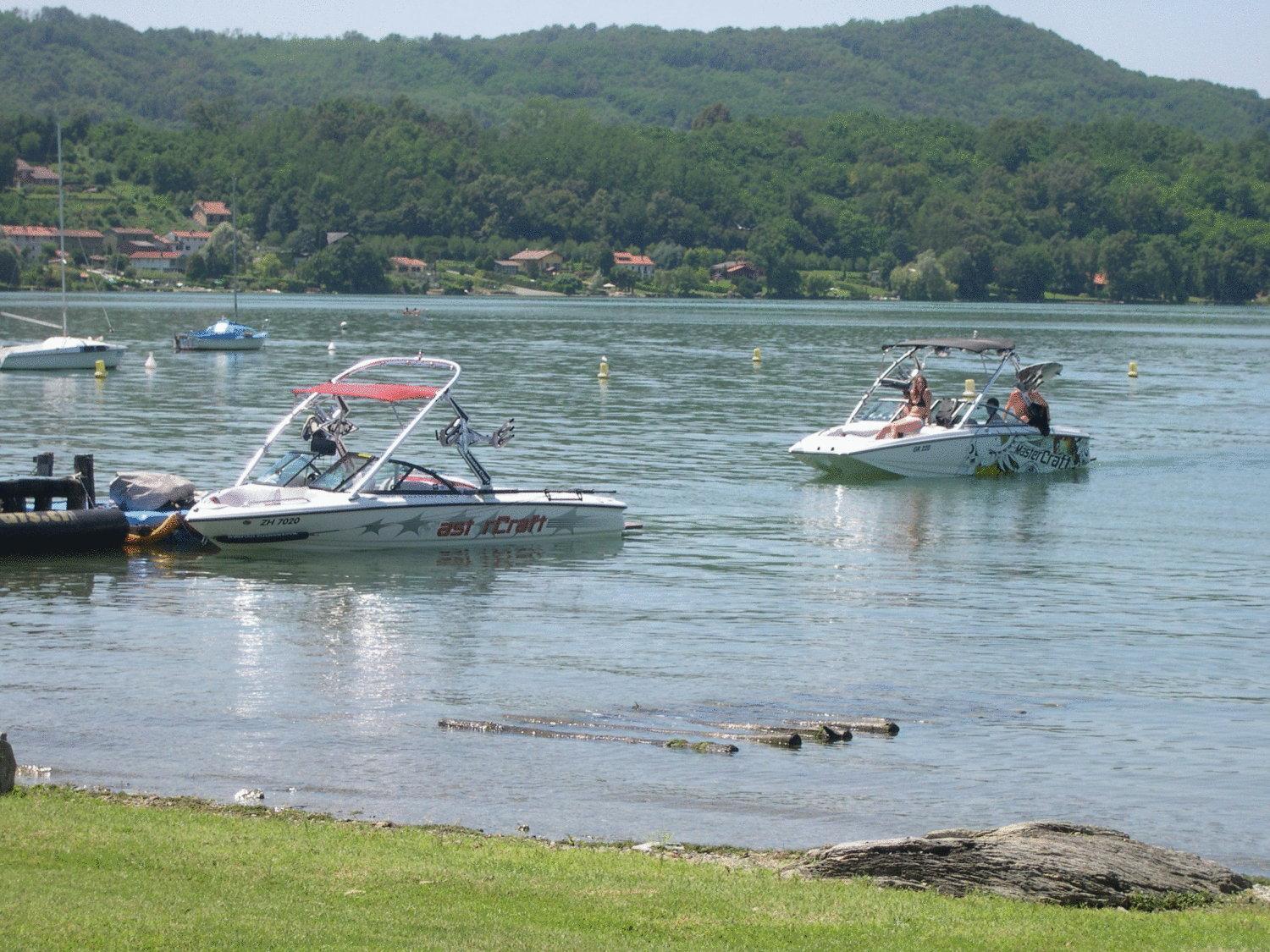 Motorboats float on calm lake water beneath green, forested hills in bright daylight.