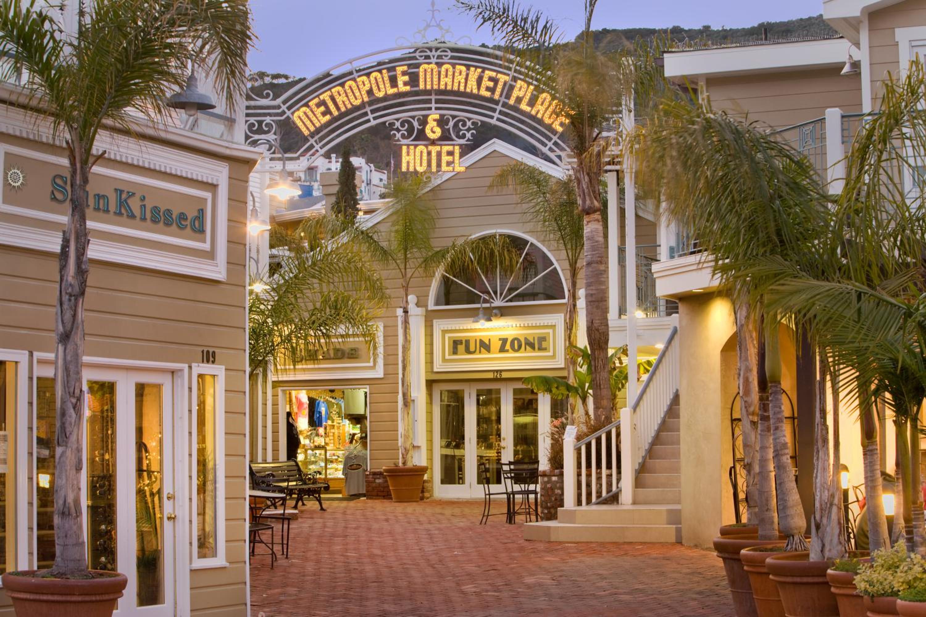 Illuminated archway framing a brick courtyard lined with shops and palm trees at twilight.