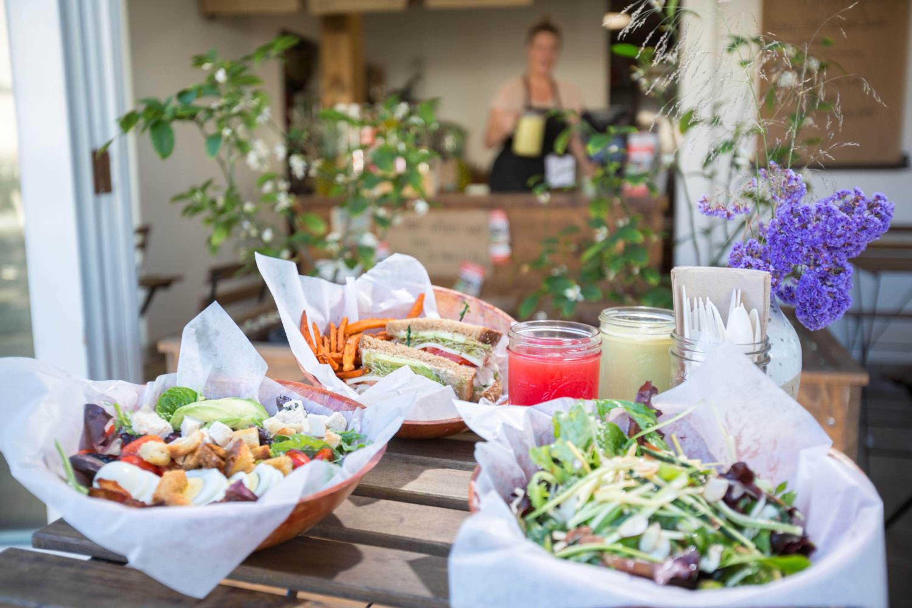Outdoor table set with salad bowls, sandwiches, and fresh juices outside a cafe counter.