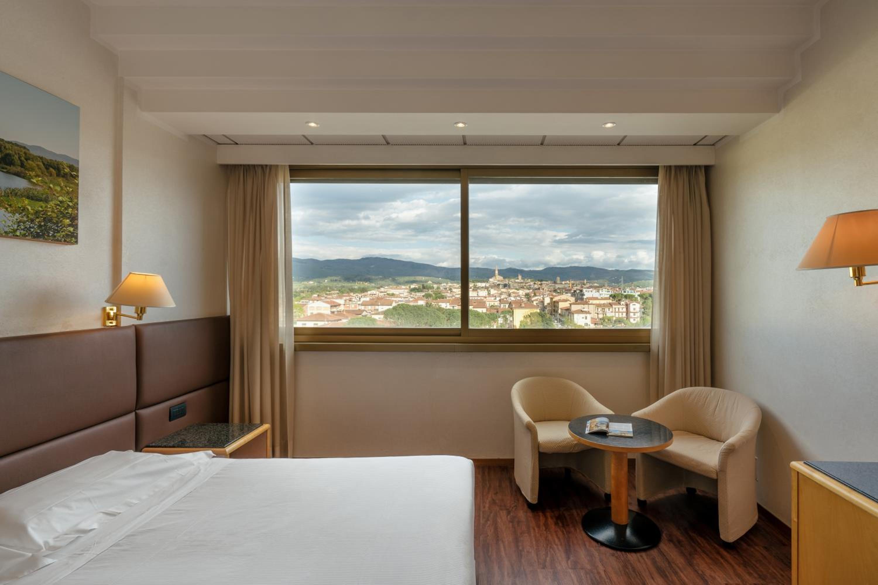 Hotel bedroom with wood flooring, cushioned armchairs, and a wide window looking out over the town.