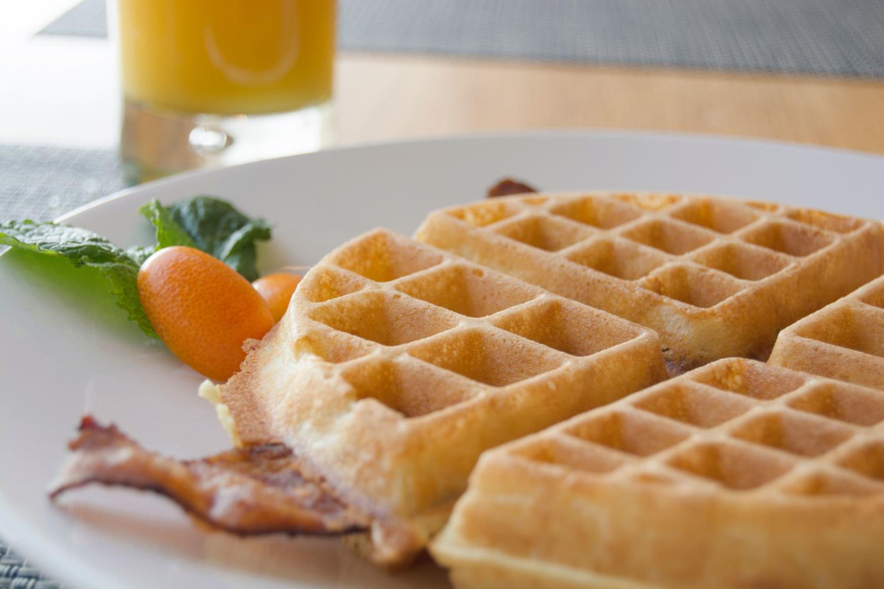 Sliced waffles served with bacon and kumquats on a white plate next to orange juice.
