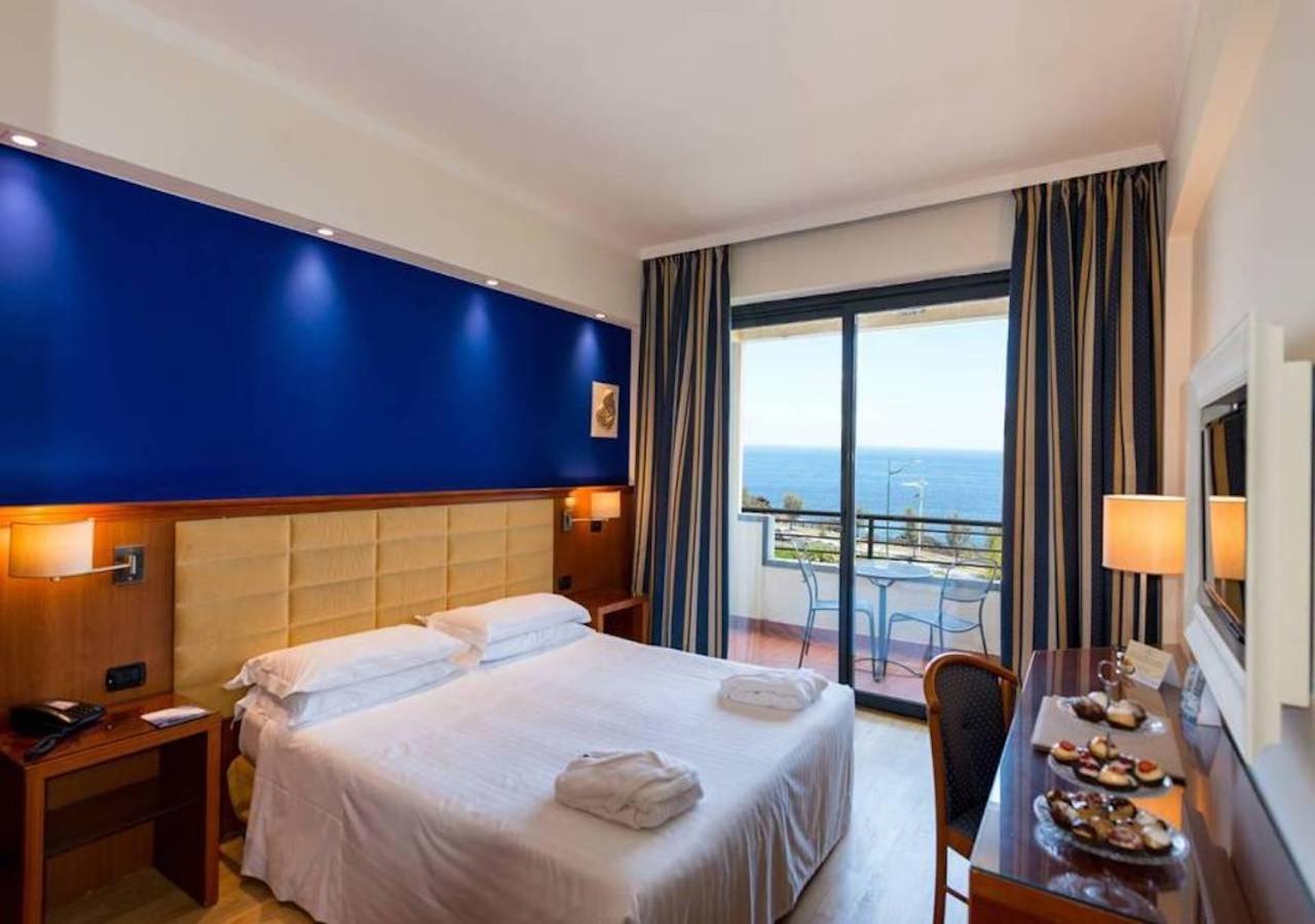 A bedroom with a blue feature wall, a cushioned headboard, and glass doors leading to a balcony overlooking the sea.