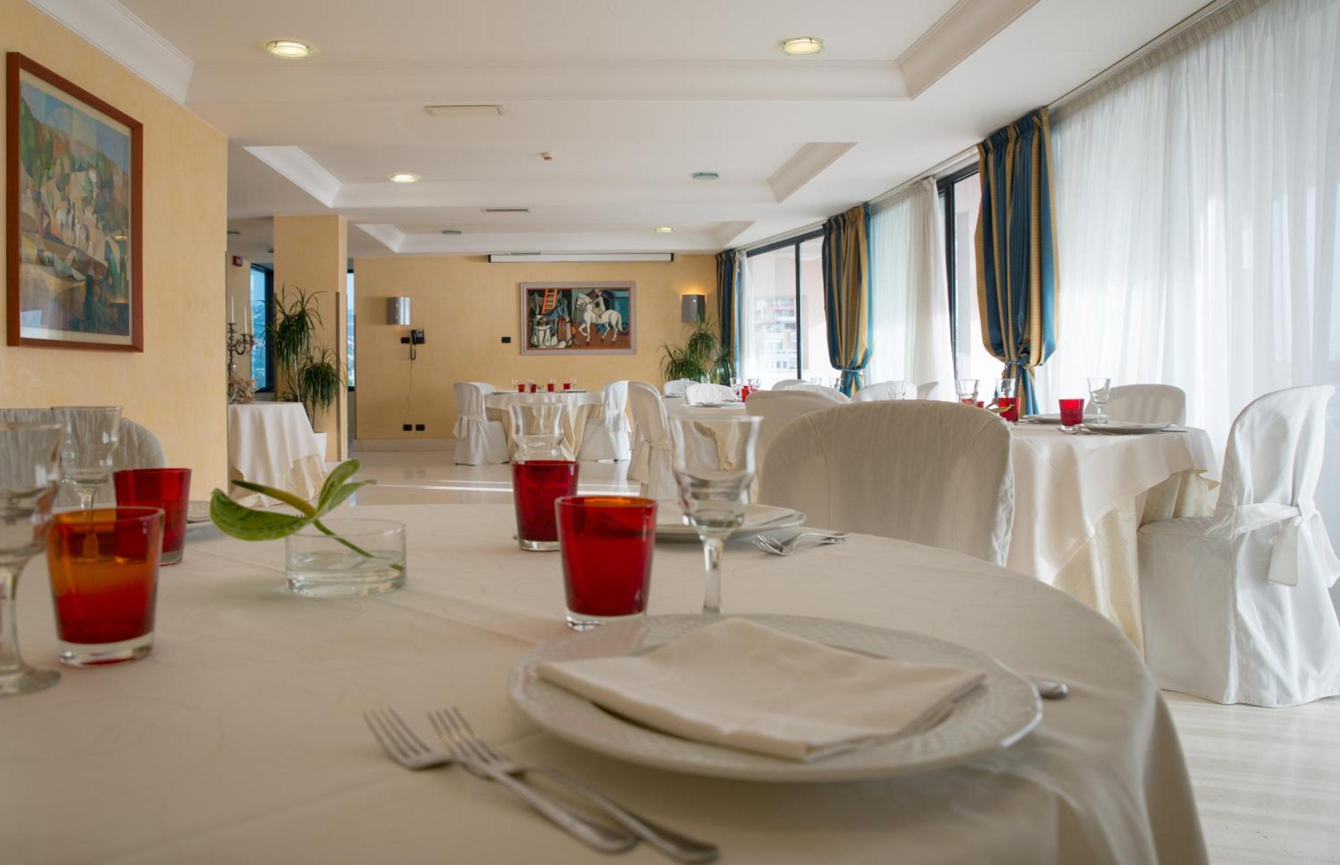 A dining room with white tablecloths, draped chairs, and red glasses set beside large windows.