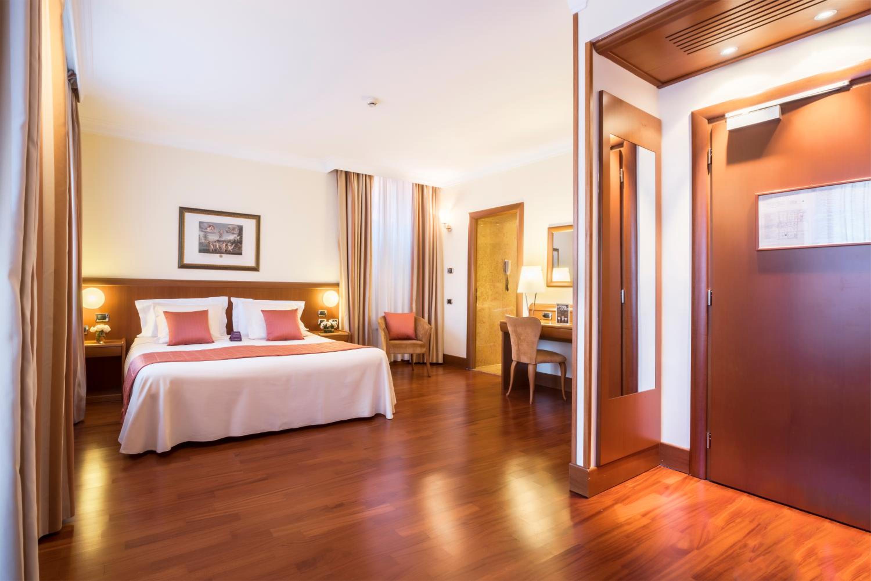 A hotel bedroom with hardwood floors, a made bed with coral cushions, and a wooden vanity desk.