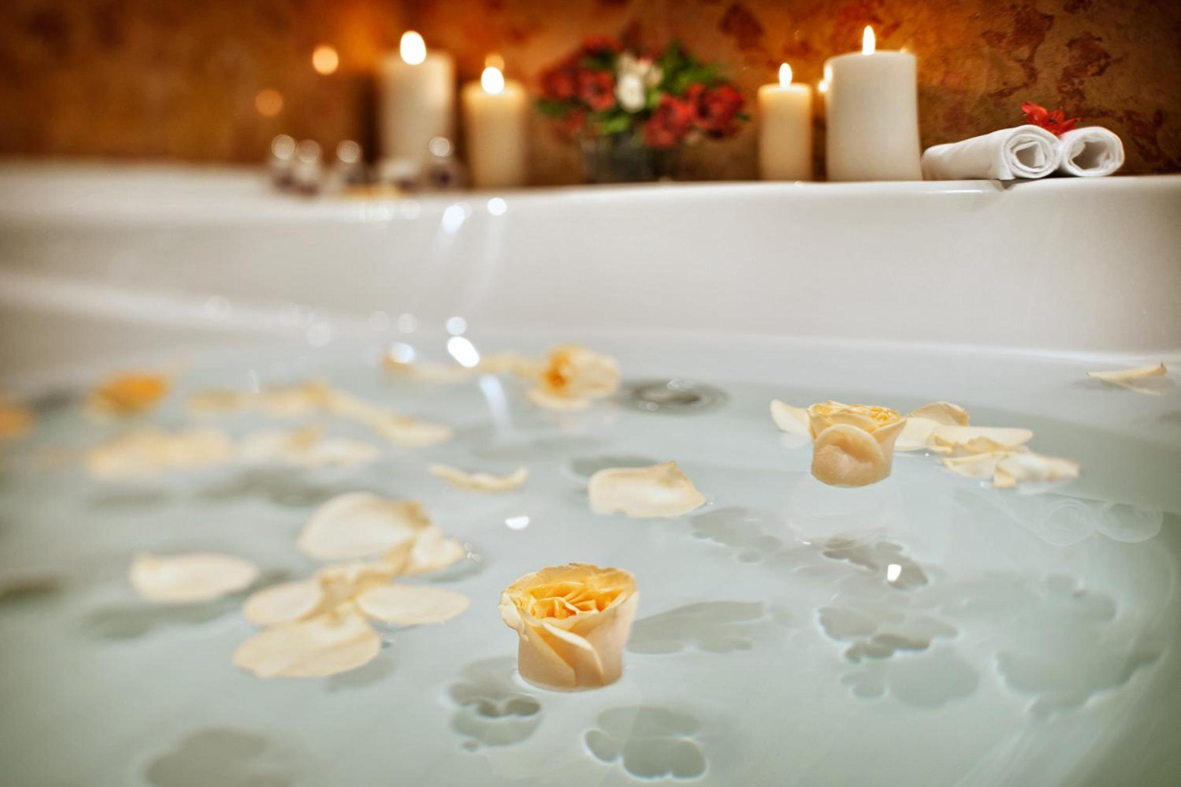 Yellow rose petals float in a white bathtub beside burning pillar candles.