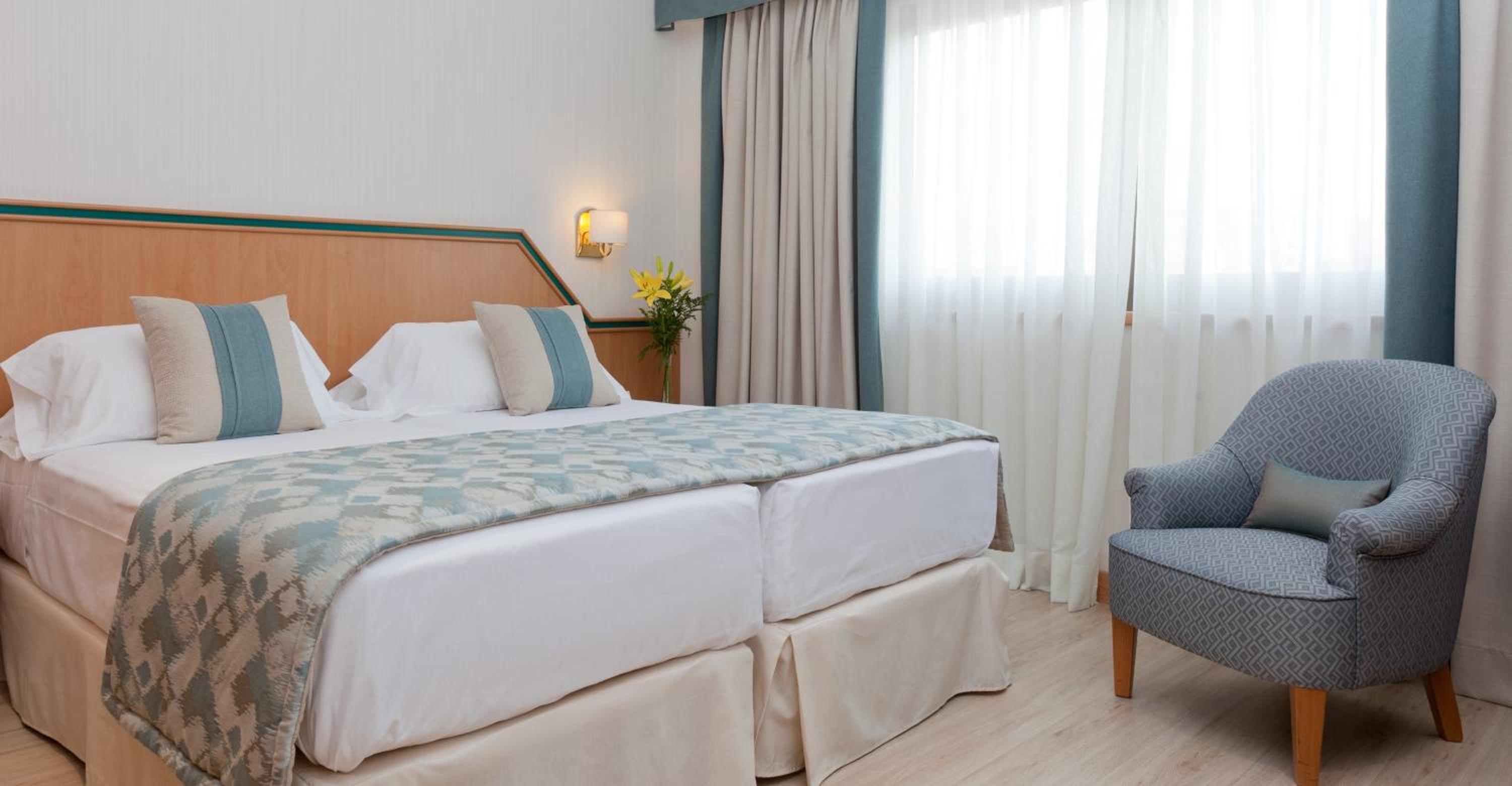 A bedroom at Hotel Praga with patterned bedding, a wooden headboard, an armchair, and a curtained window.