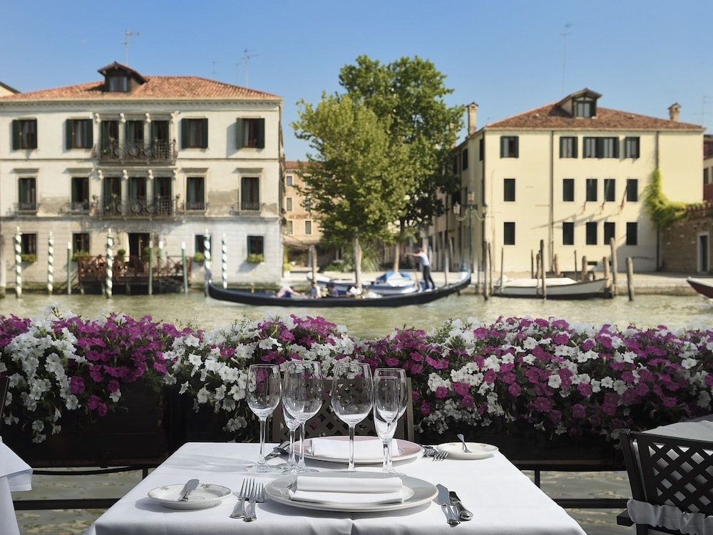 A dining table overlooks a canal with passing gondolas beyond blooming purple petunias.