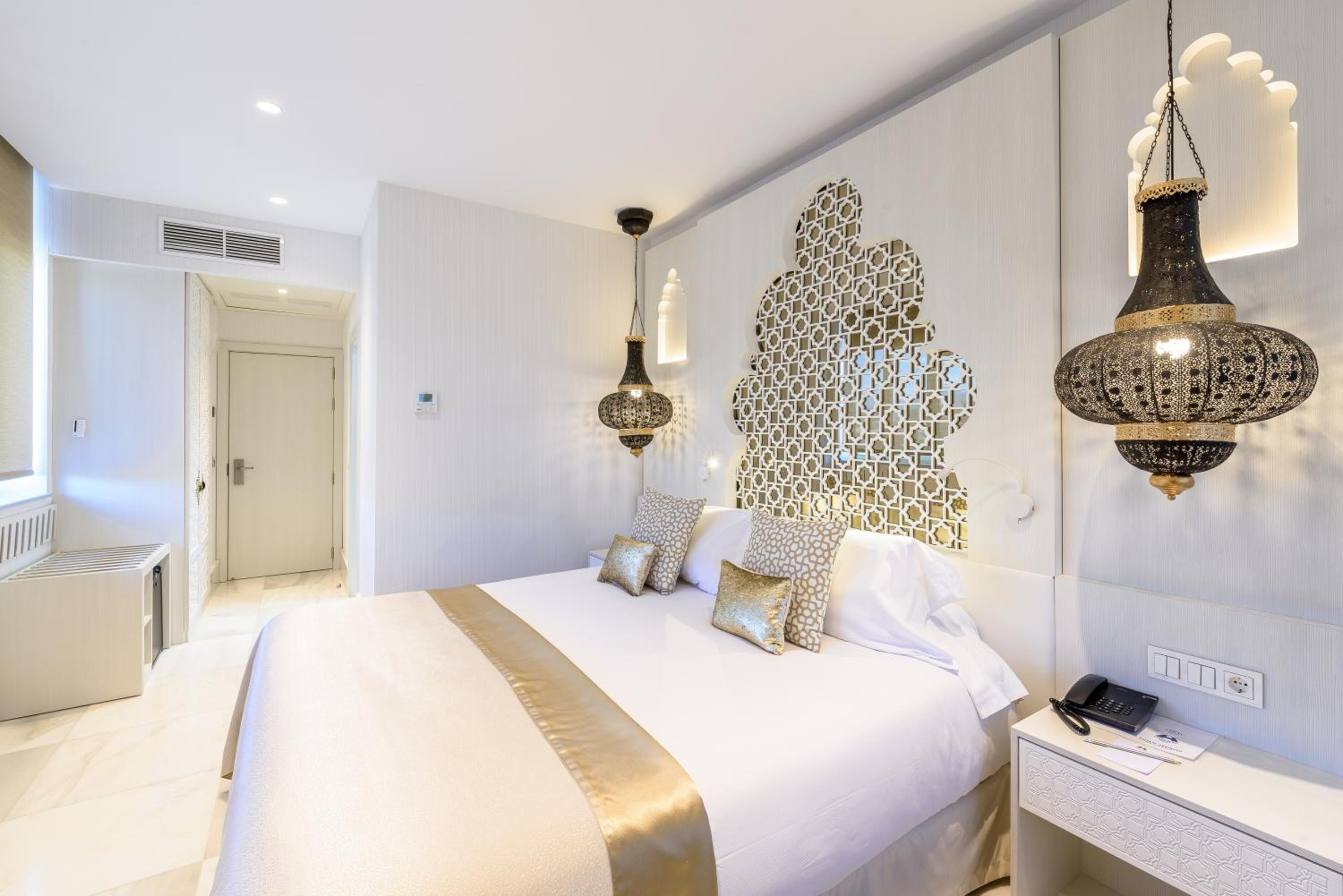 A bright hotel bedroom with an ornate geometric headboard panel and hanging pierced metal lanterns.