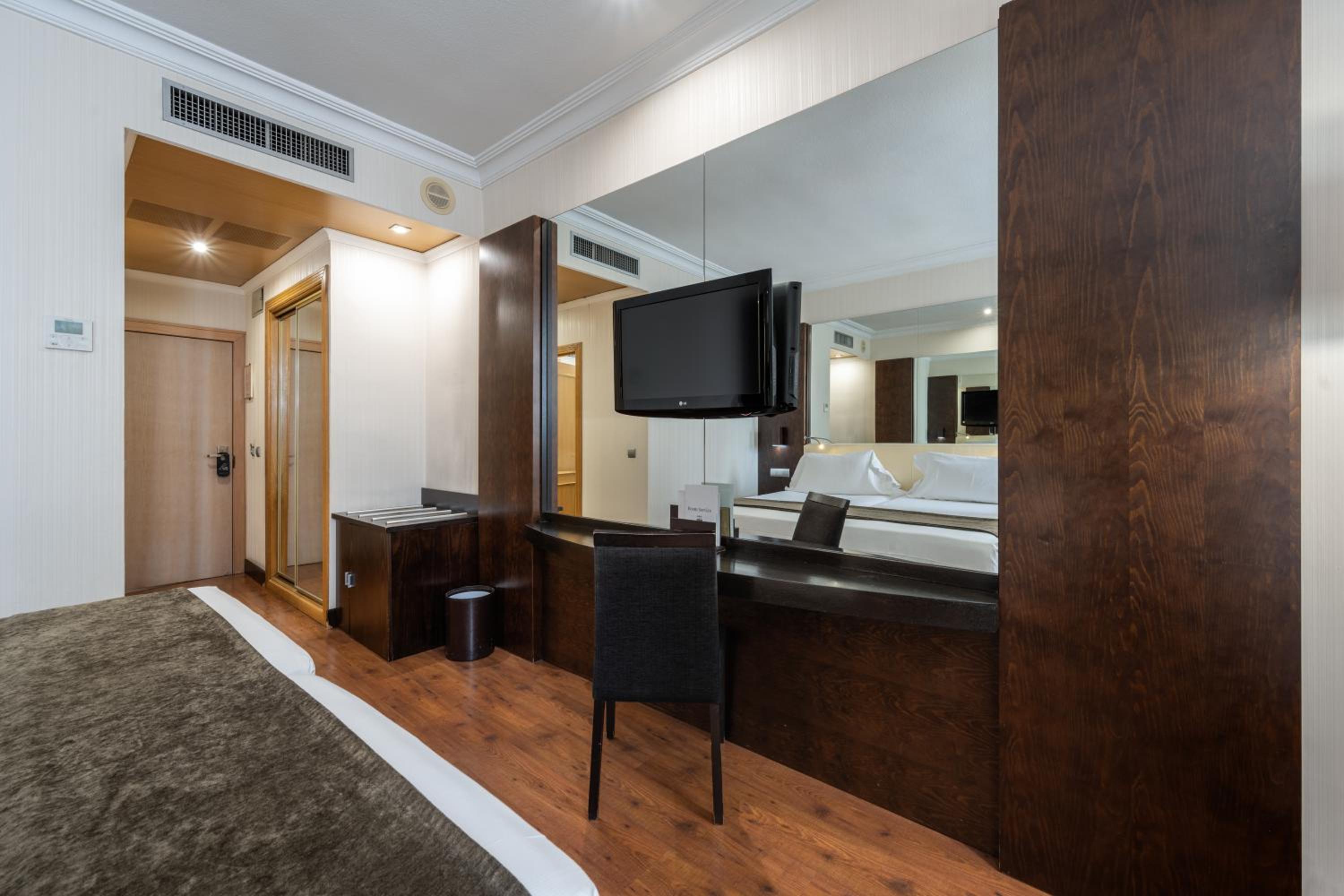 A hotel bedroom corner featuring a dark wood desk with a chair, a large mirror, and a wall-mounted TV.