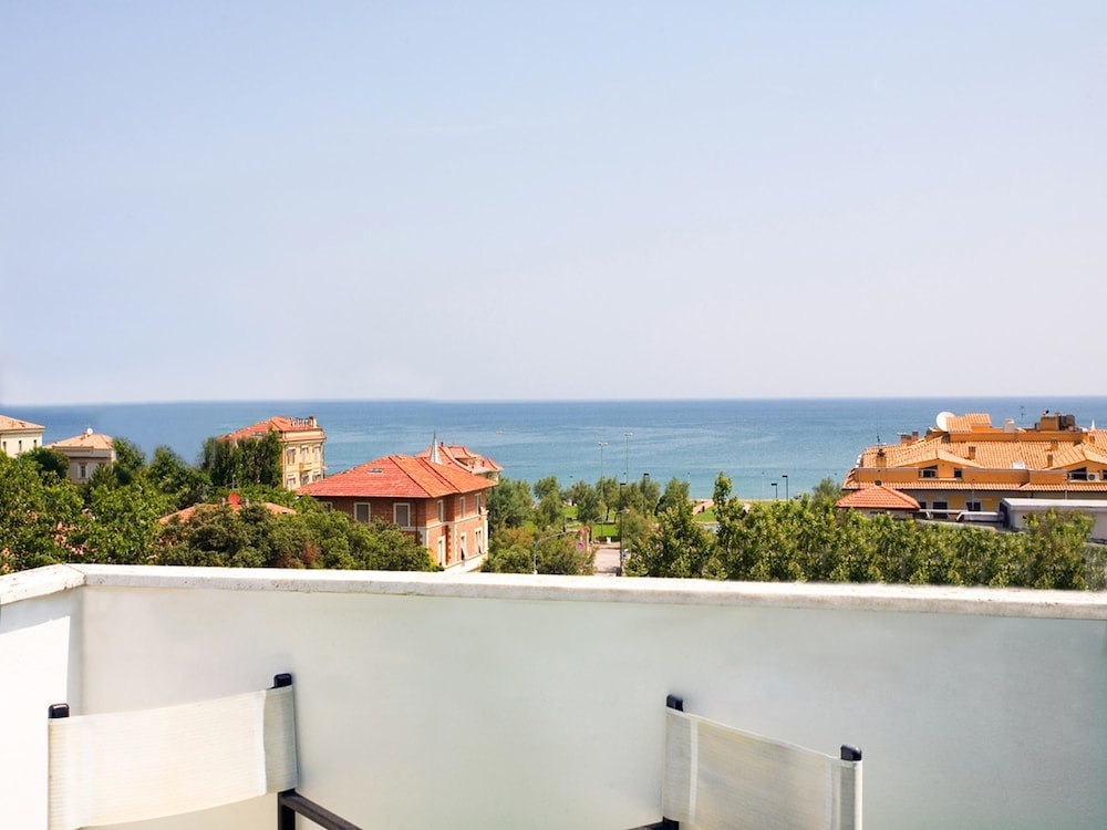 A balcony view overlooks terracotta rooftops and green trees stretching toward the calm blue sea.