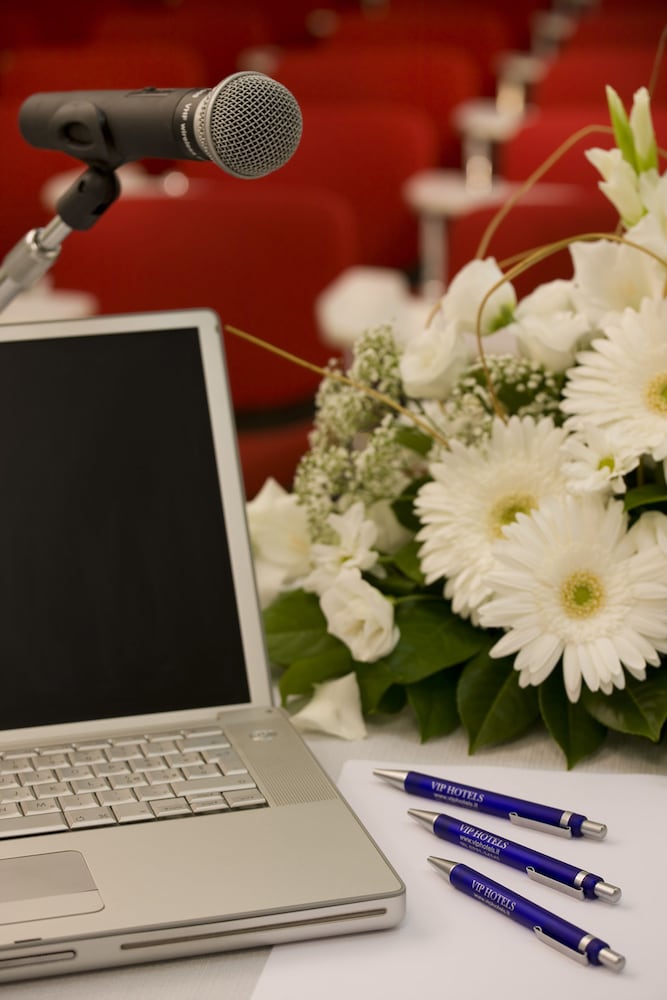 A presentation table holds an open laptop, a microphone, branded blue pens, and a bouquet of white flowers.