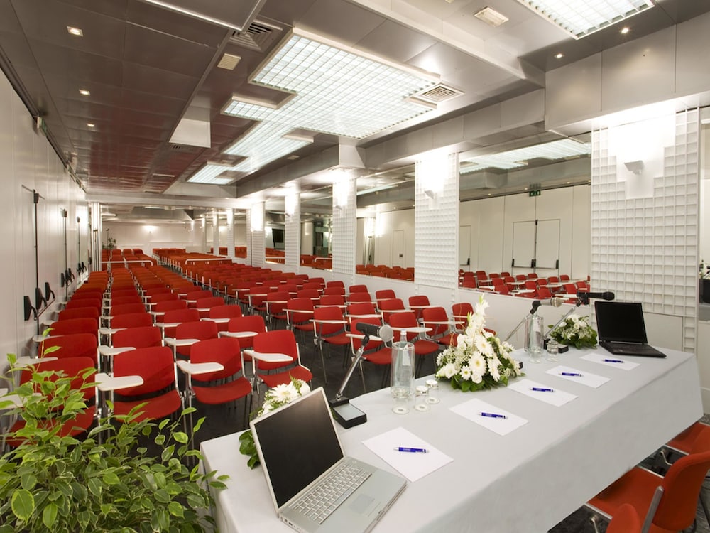 Hotel Savoy conference hall features rows of red chairs facing a draped table with laptops and microphones.