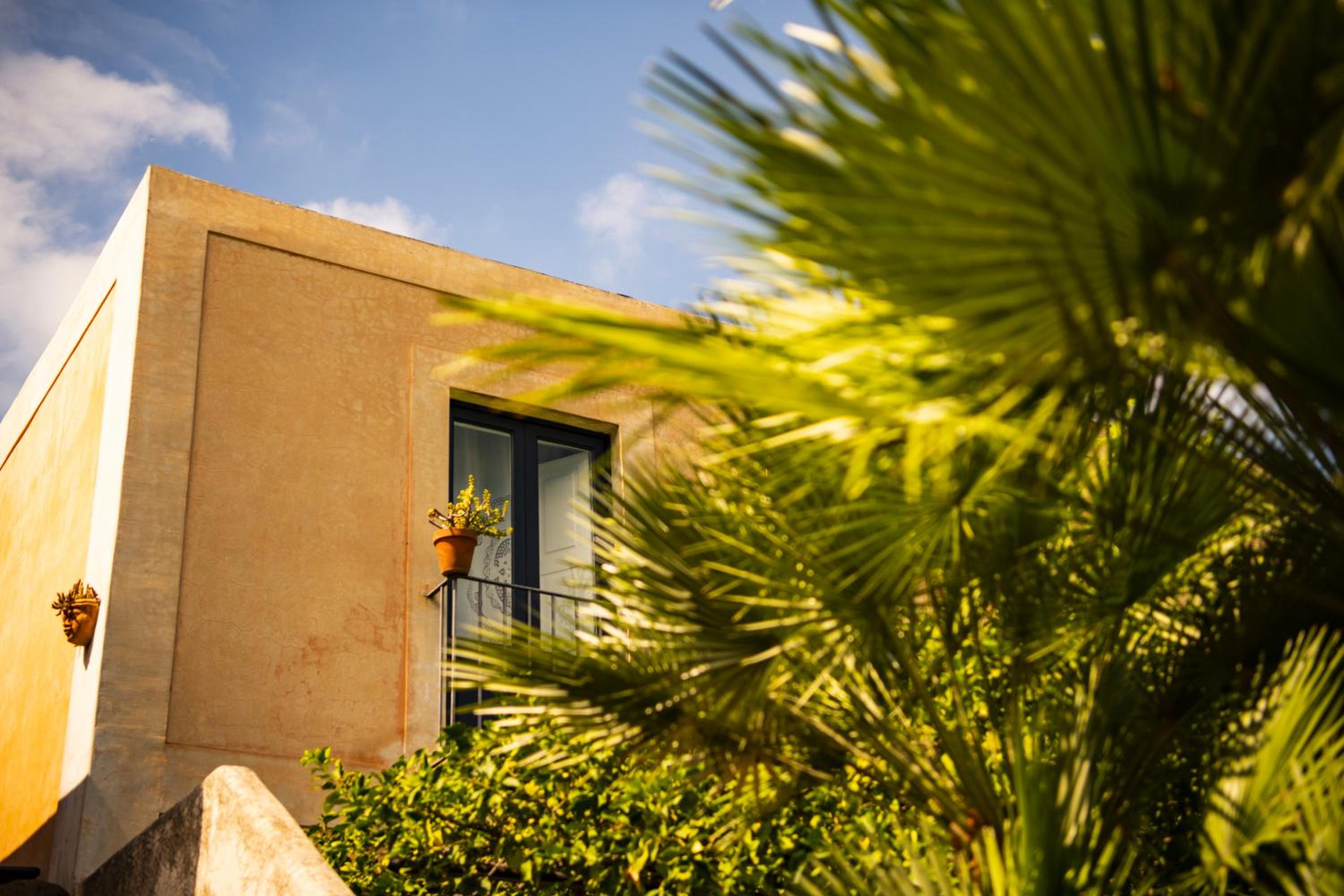 Sunlit ochre facade with a small balcony and potted plant, framed by green palm fronds.