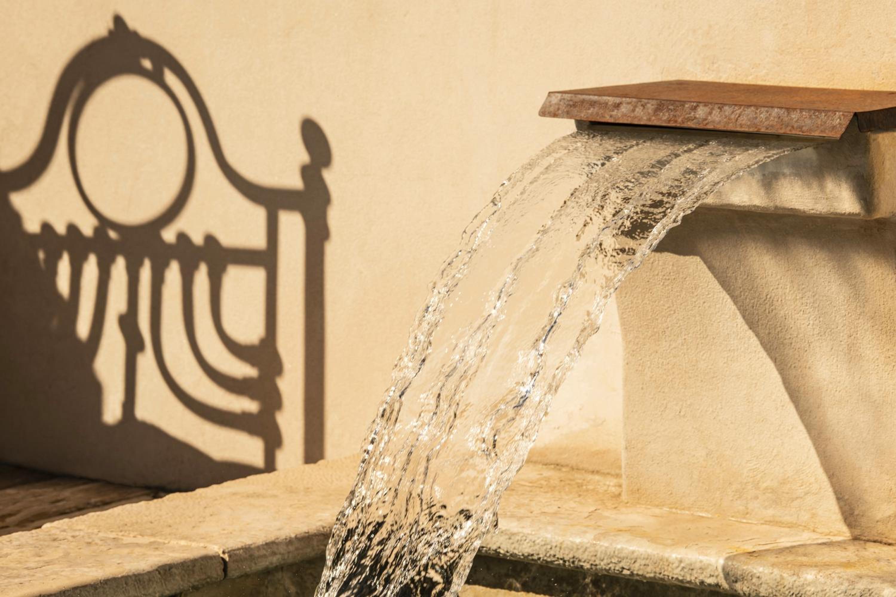 Water cascades from a stone fountain spout, casting shadows of ornate ironwork onto a sunlit wall.