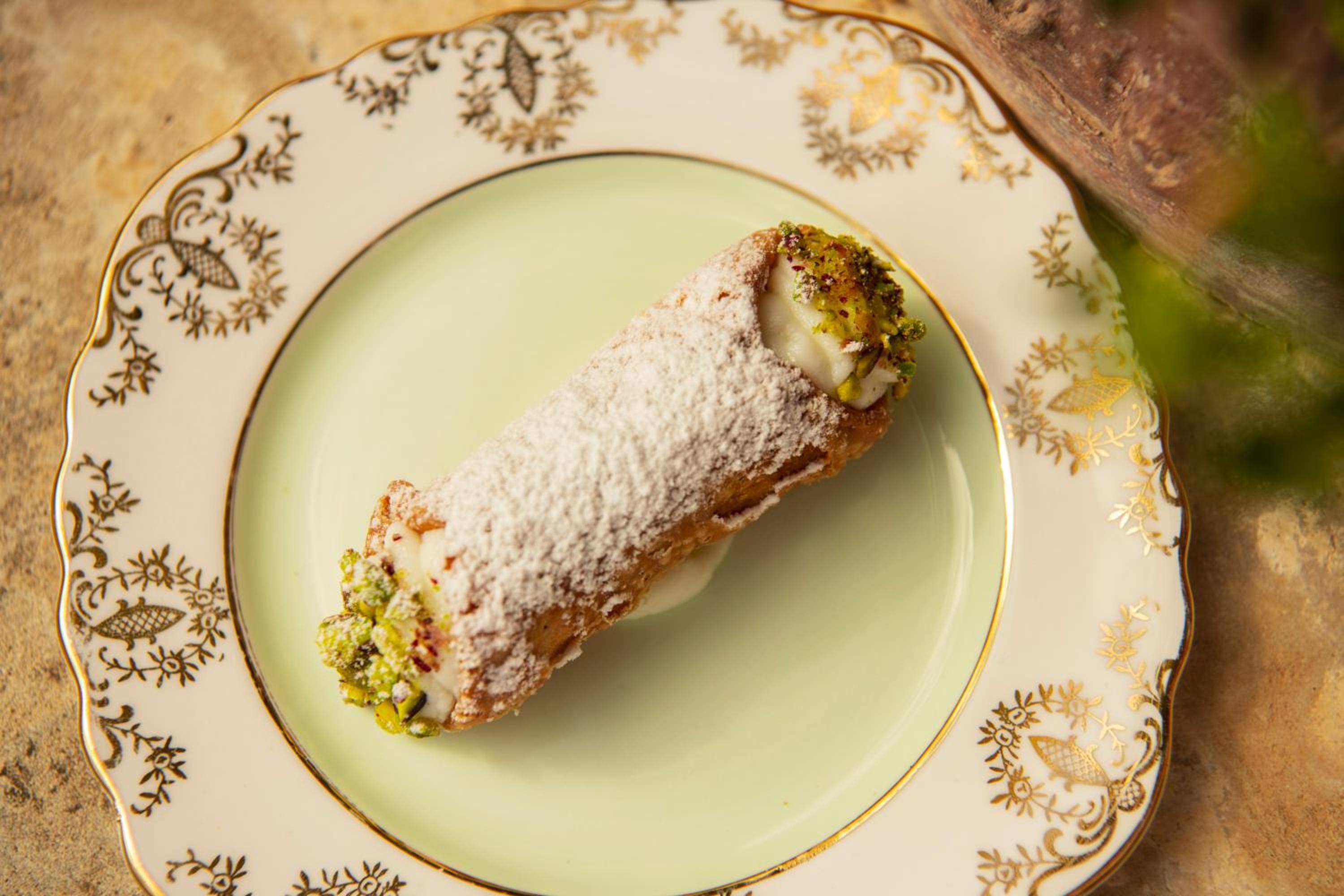 A cannolo dusted with powdered sugar and chopped pistachios on a vintage porcelain plate with gold trim.