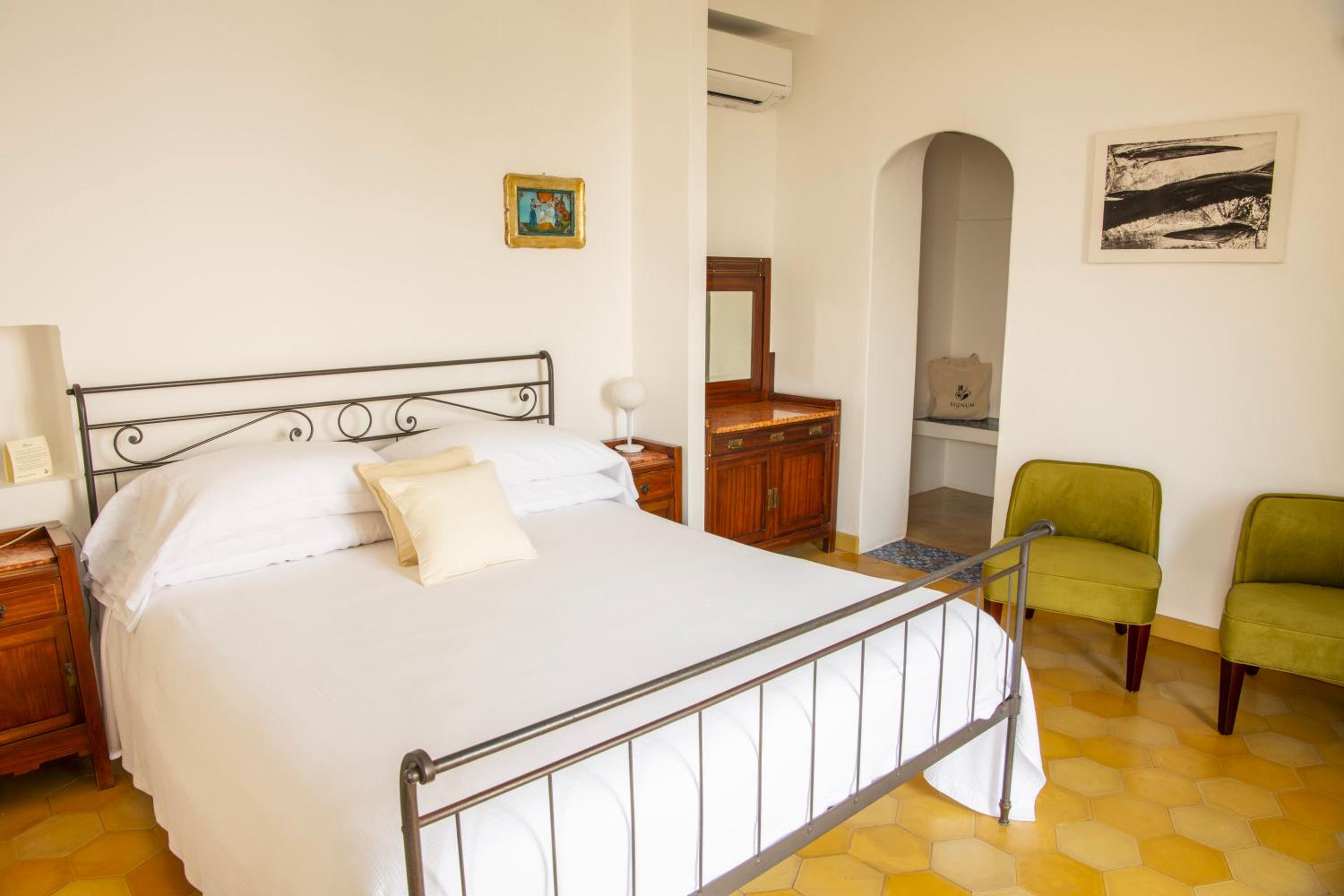 A bedroom at Hotel Signum with a wrought-iron bed, yellow tiled floor, green armchairs, and an arched doorway.