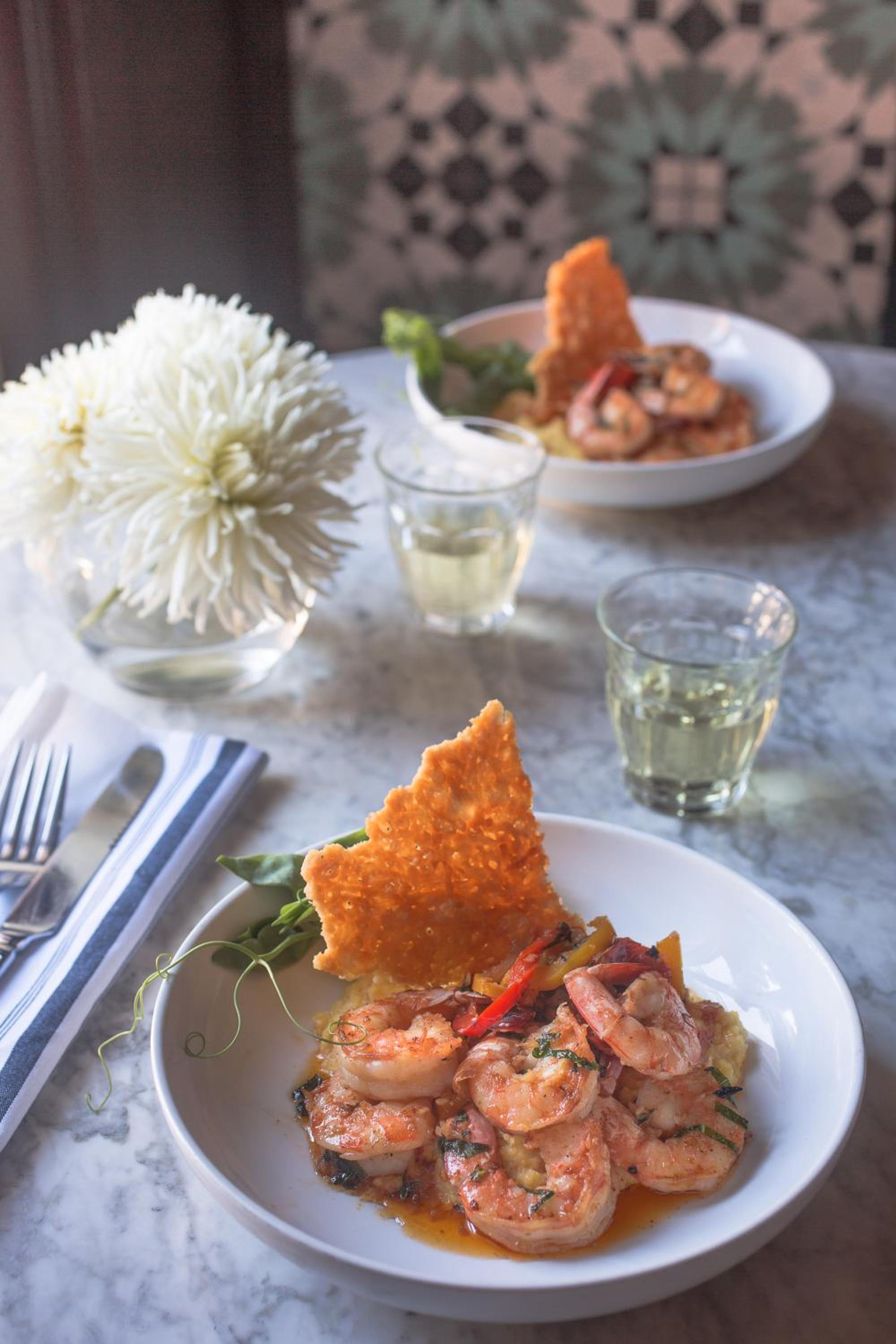 Dishes of shrimp with crisp garnish and wine glasses beside a vase of white flowers on a marble table.