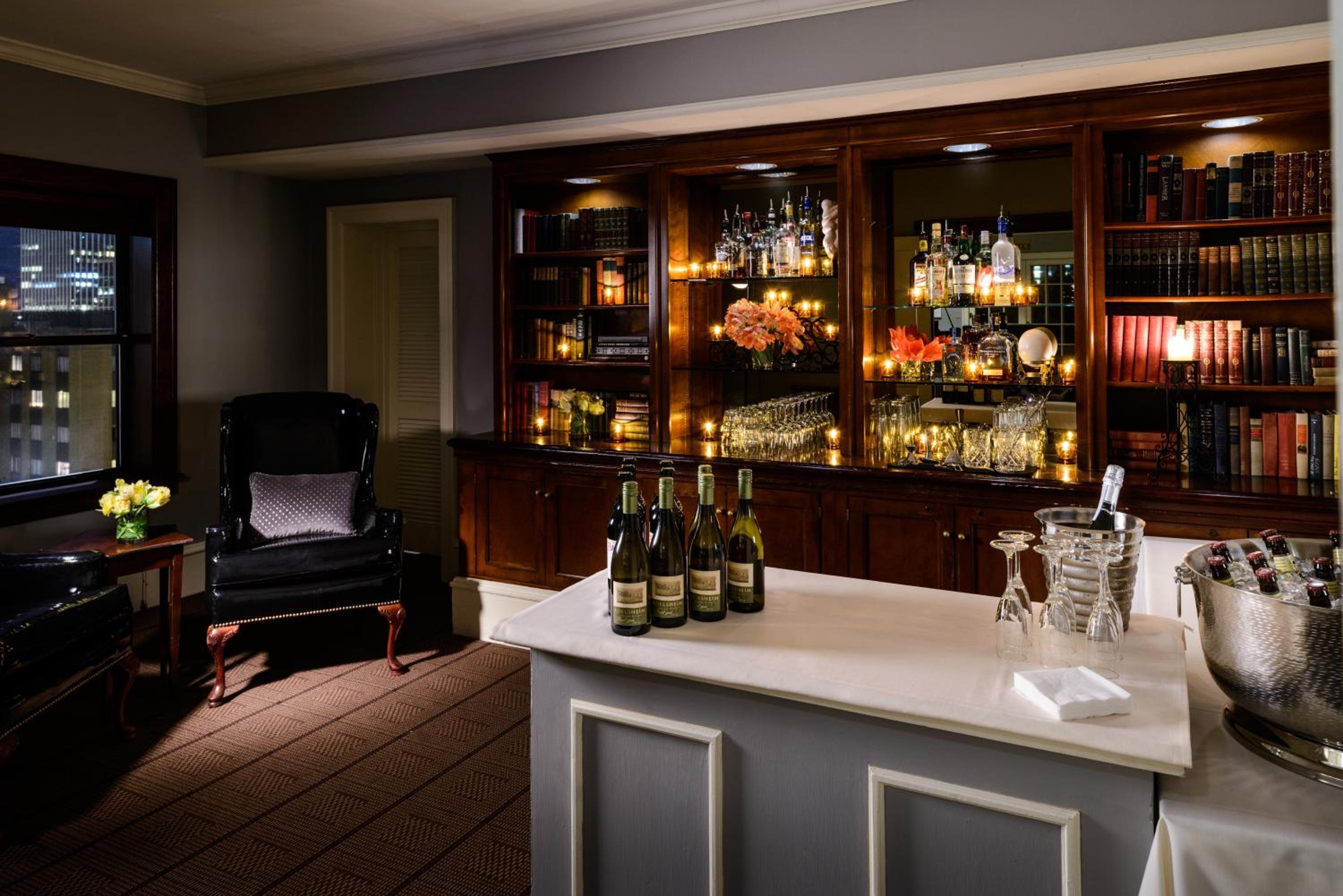 A dark wood backbar with lit shelves and books, facing a serving counter set with wine bottles and glassware.