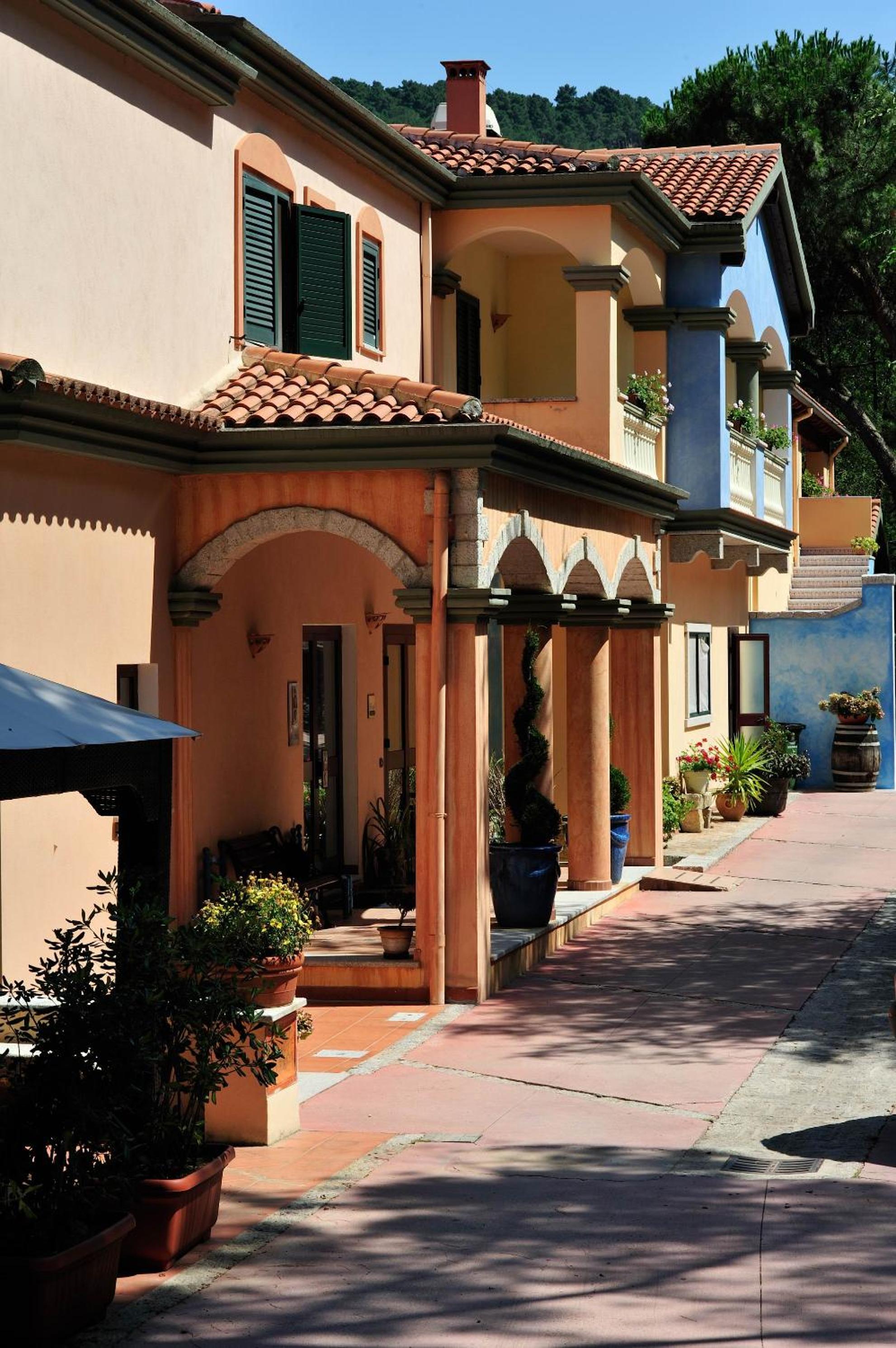 Facade of Hotel Su Lithu with terracotta walls, tiled roofs, and an arched ground-floor colonnade in bright sunlight.