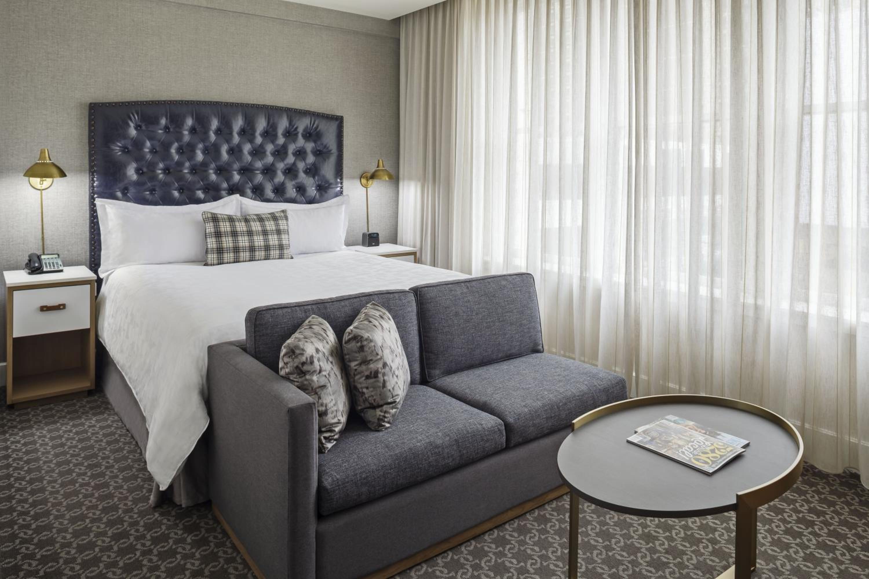 A hotel bedroom with a tufted leather headboard, a gray upholstered settee, and sheer window curtains.