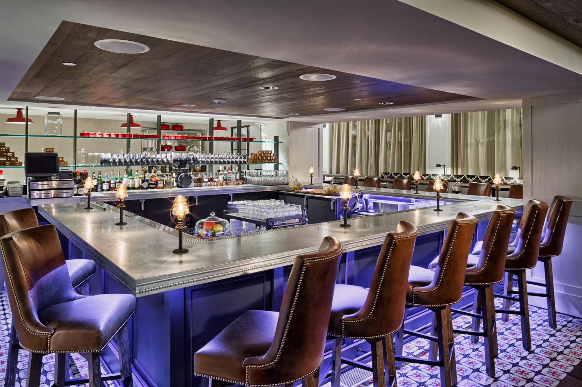 A wrap-around metal bar counter with leather barstools, mood lighting, and patterned floor tile.