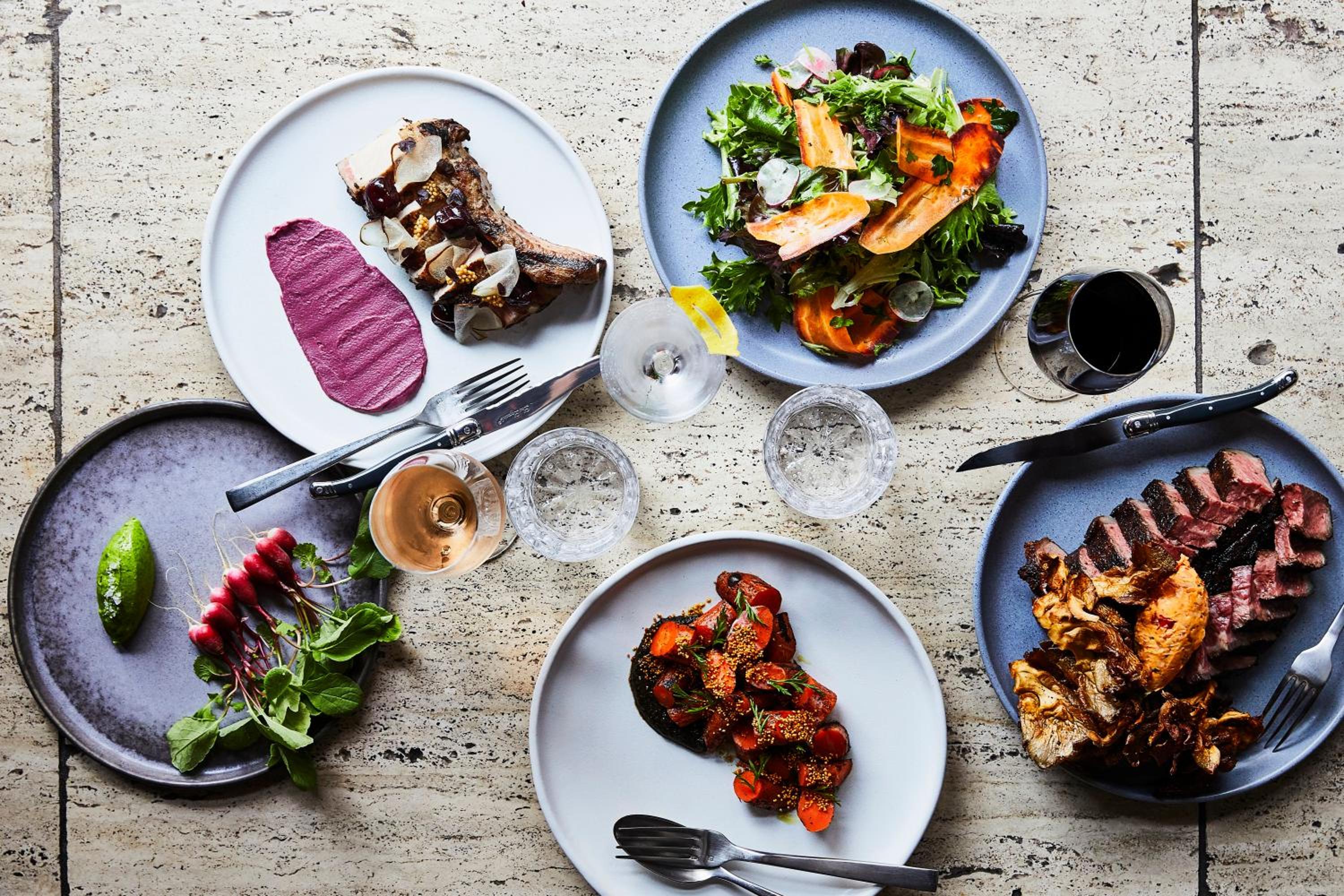 Overhead view of roasted meats, salad, carrots, and glasses of wine set on a stone tabletop.