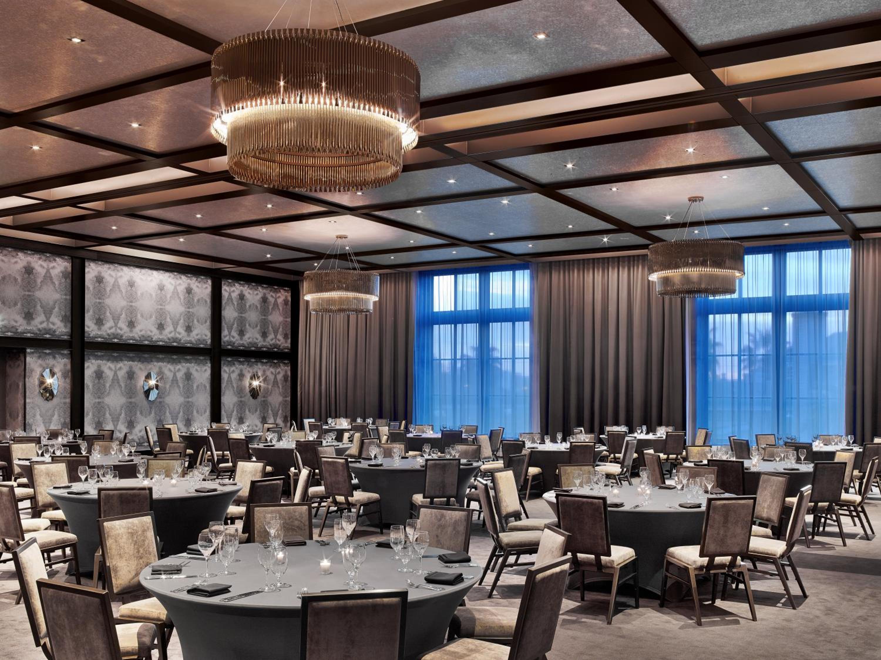 A ballroom with a coffered ceiling, hanging chandeliers, and round tables set for dining before tall windows.