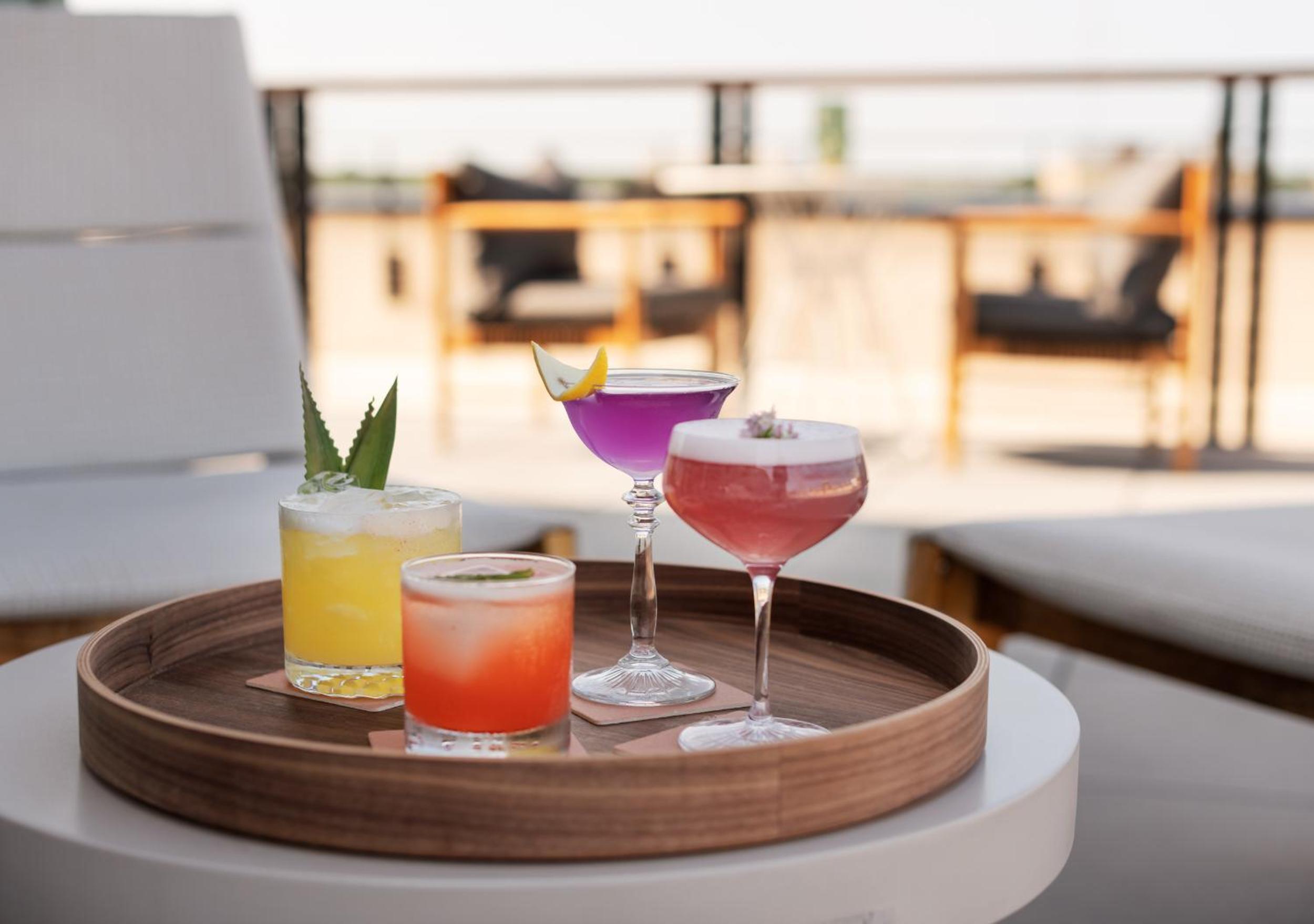 Colorful cocktails served on a wooden tray on a sunny outdoor terrace.