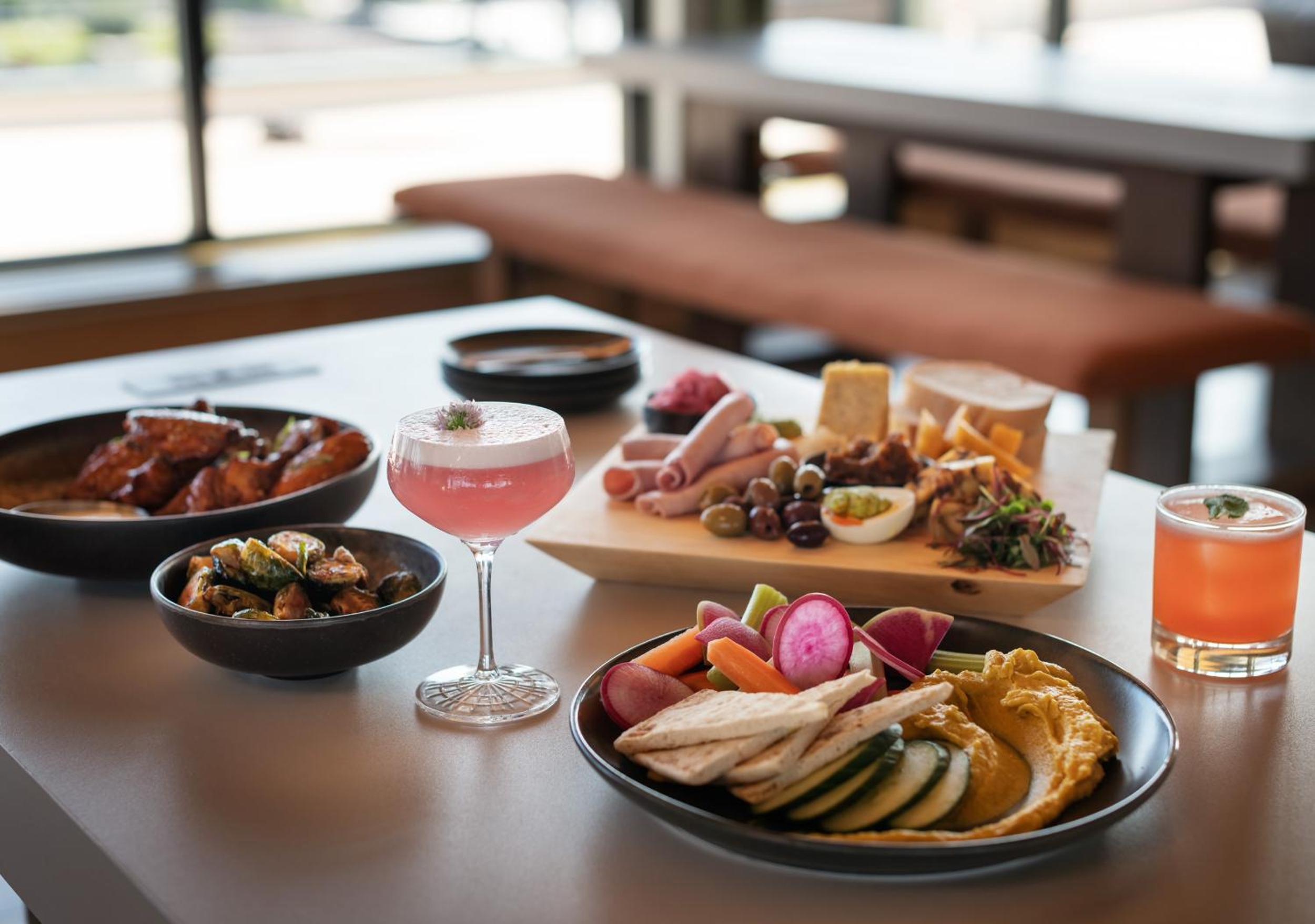 A dining table set with cocktails, hummus with pita and vegetables, charcuterie, and small plates.