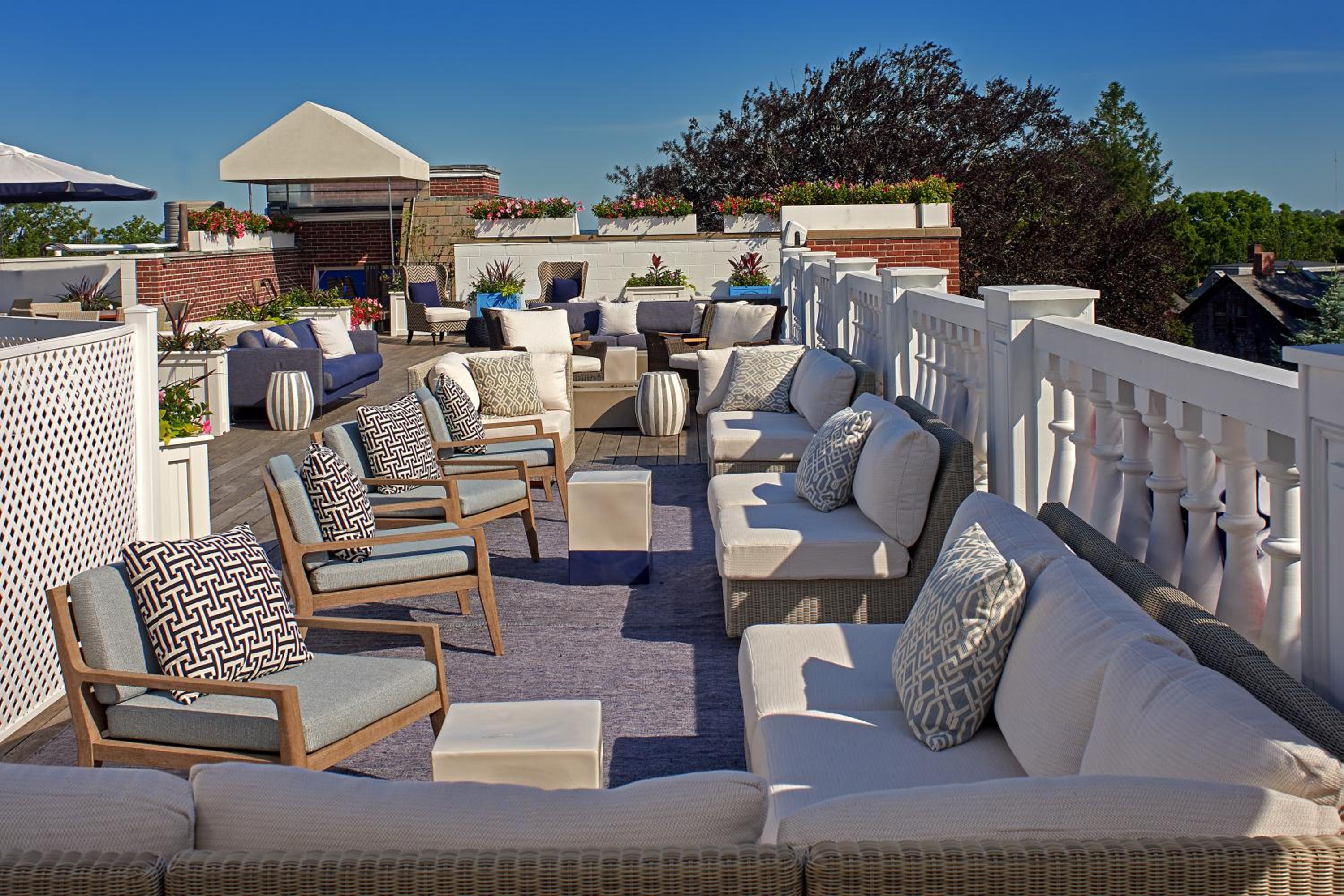 A sunny rooftop terrace furnished with cushioned wicker sofas, armchairs, and planters along a white balustrade.