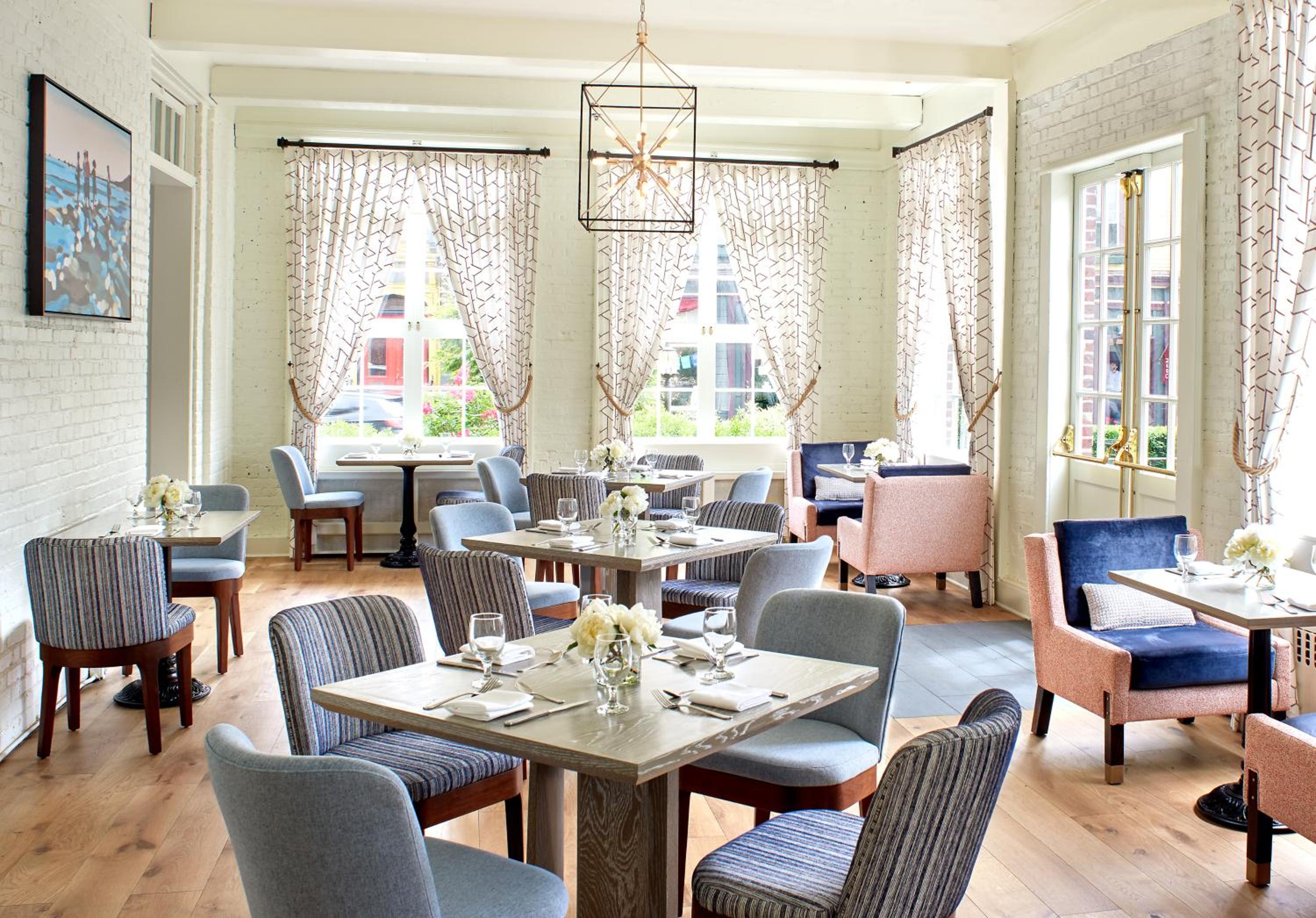 A sunlit dining room featuring whitewashed brick walls, draped windows, and tables with upholstered chairs.