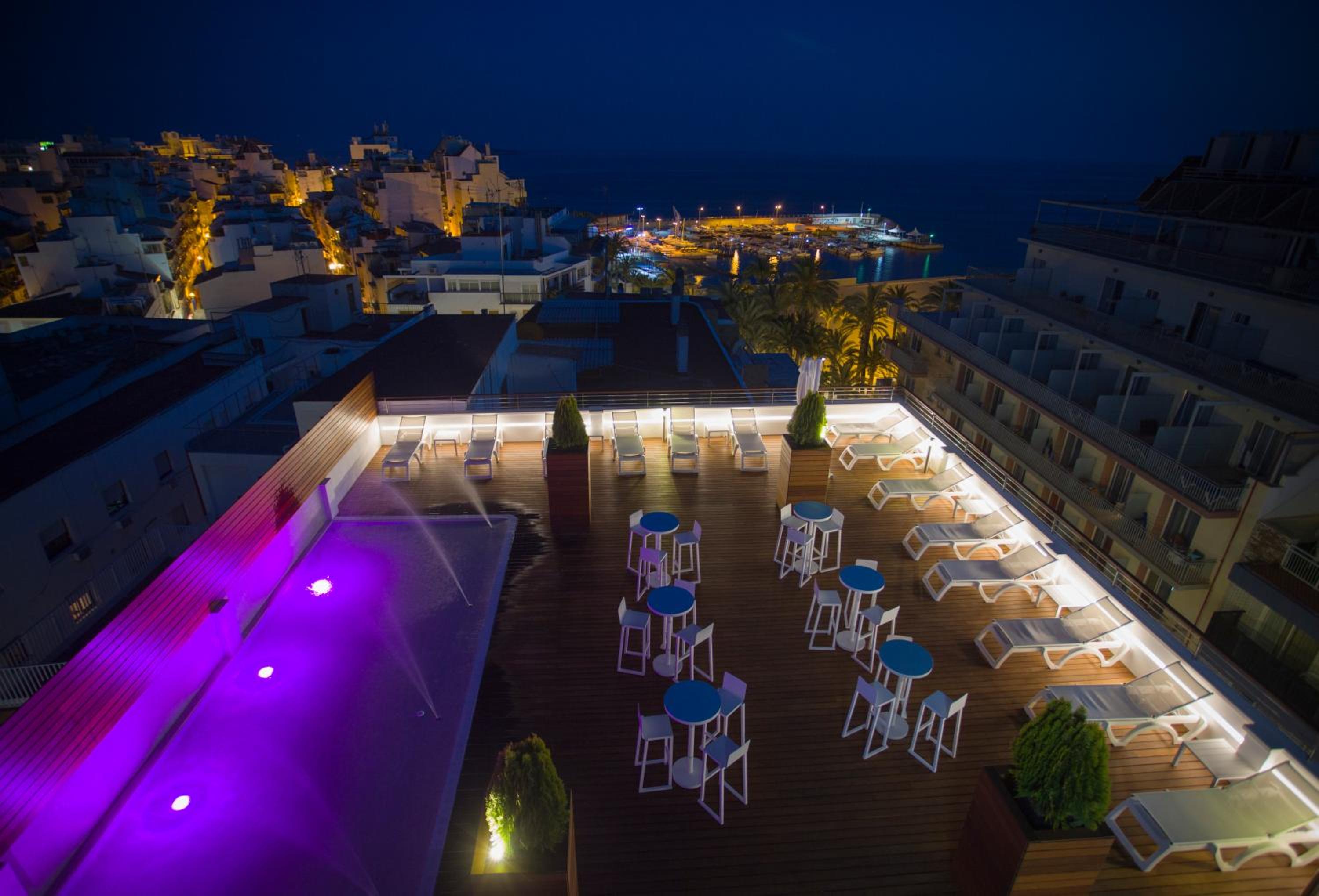 Rooftop terrace at night with illuminated water jets, high tables, sun loungers, and distant harbor lights.