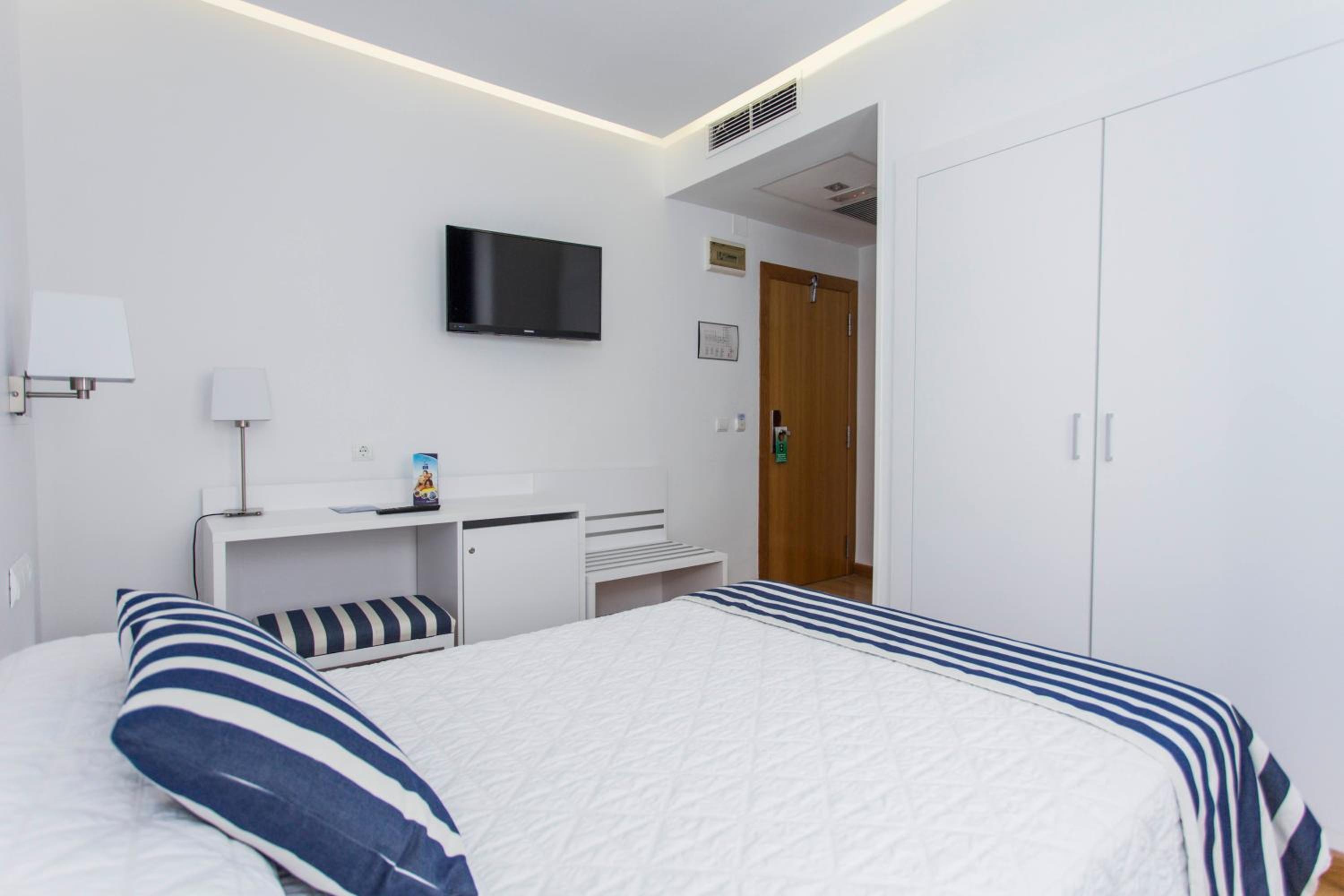 Hotel bedroom with a white and blue-striped bed, wall-mounted television, white desk, and closet.