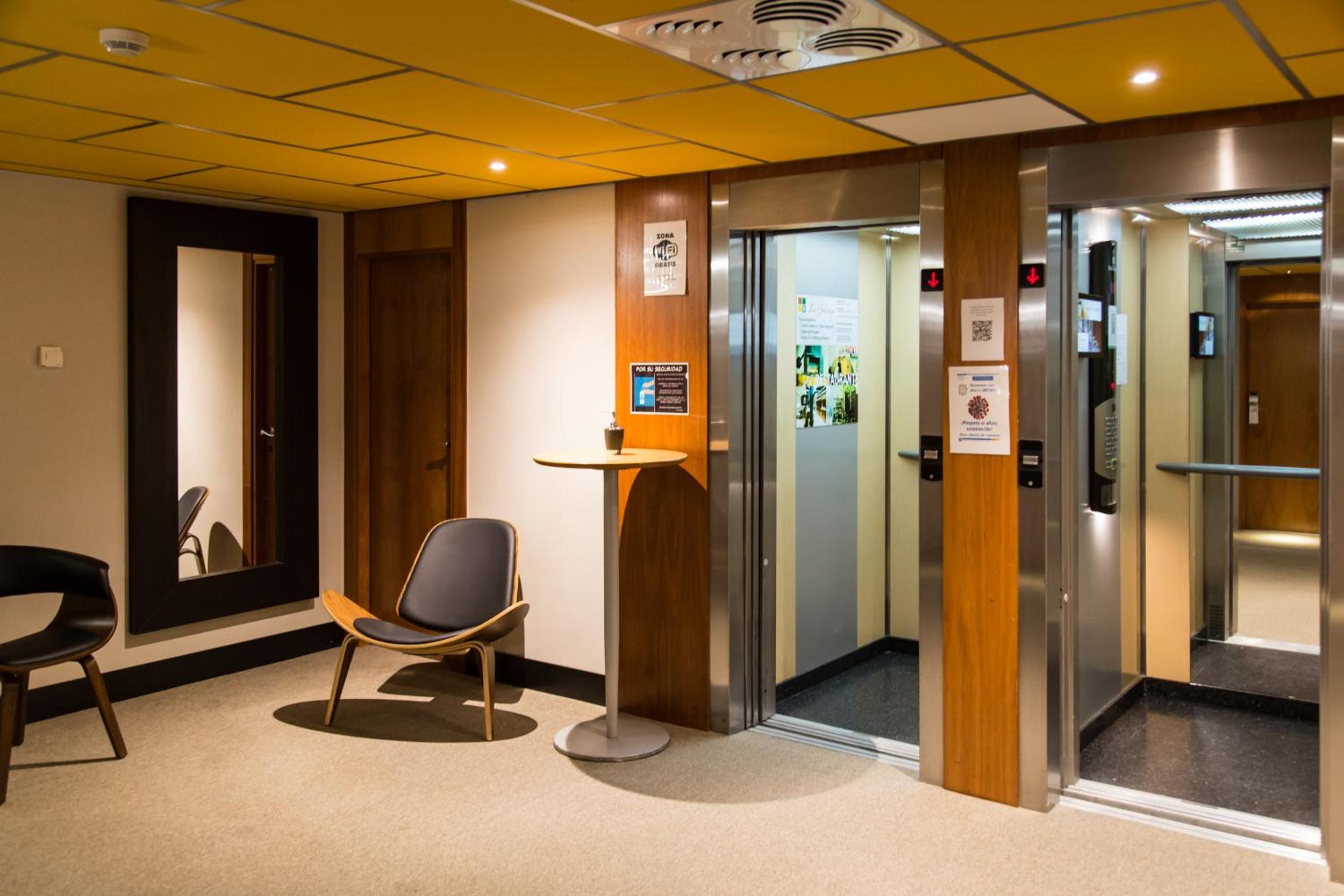 An elevator lobby featuring wood paneling, a yellow tiled ceiling, an armchair, and open elevator doors.