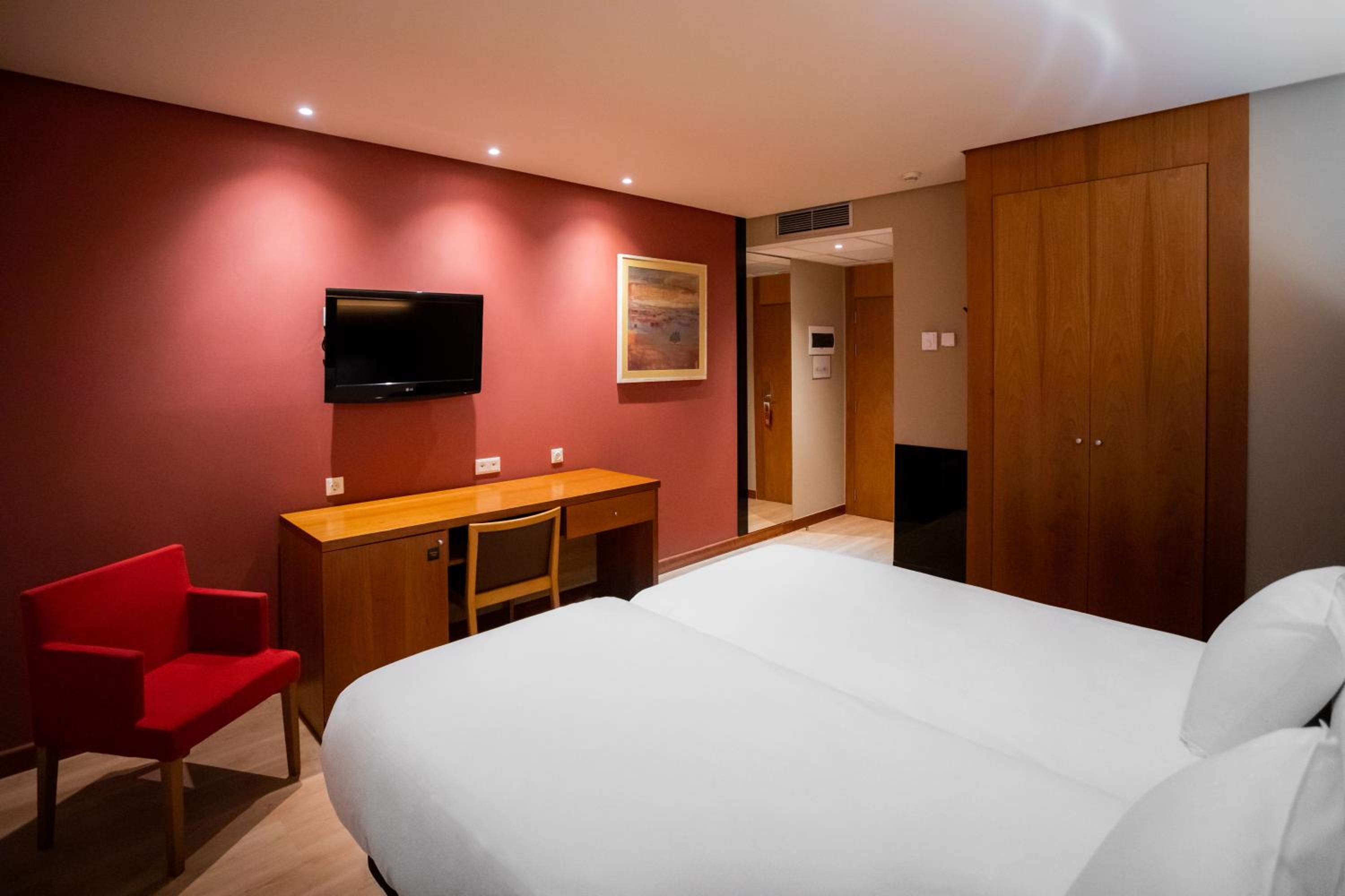 A hotel bedroom with white beds, a red accent wall, a wooden desk with a chair, and a mounted television.