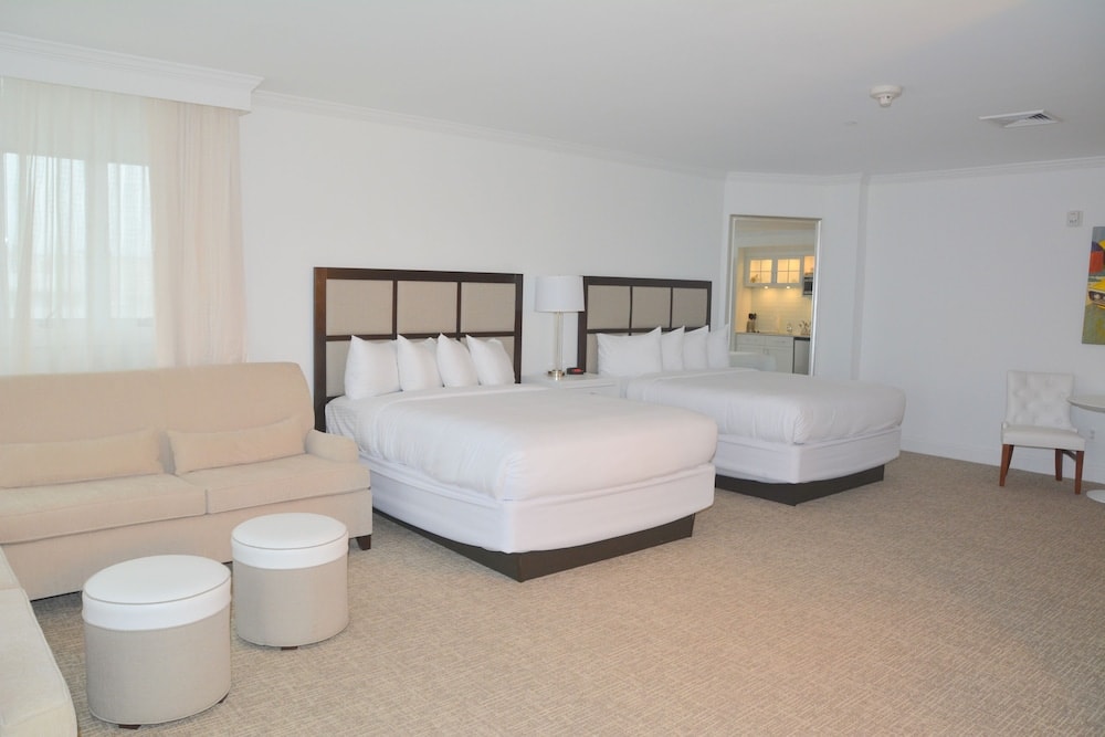Hotel bedroom with white linens on beds, matching padded headboards, beige sofa, and low cylindrical ottomans.