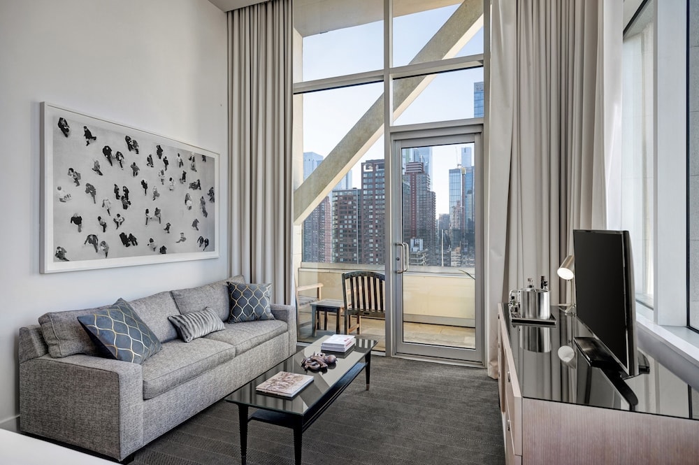 A sitting room with a grey sofa, glass coffee table, and tall windows opening to a balcony with city views.