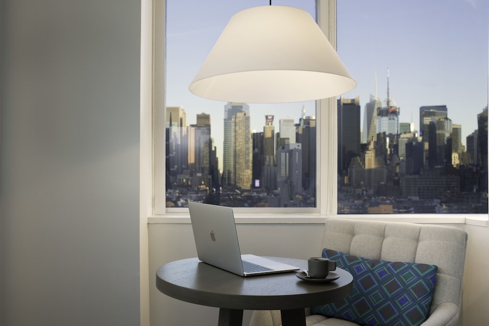 A round wooden table with a laptop and coffee cup beside a window overlooking Manhattan skyscrapers.