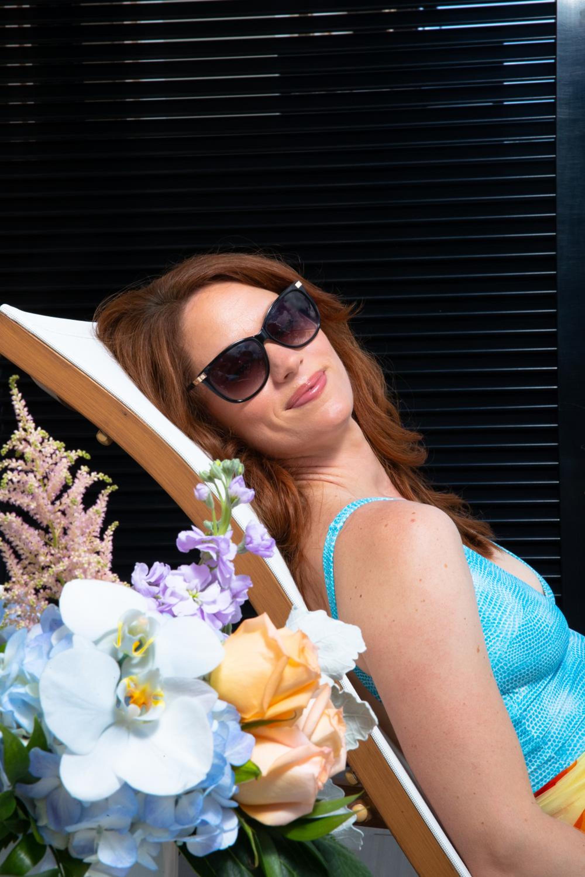 A woman wearing sunglasses relaxes on a cushioned lounger beside a vibrant floral bouquet.