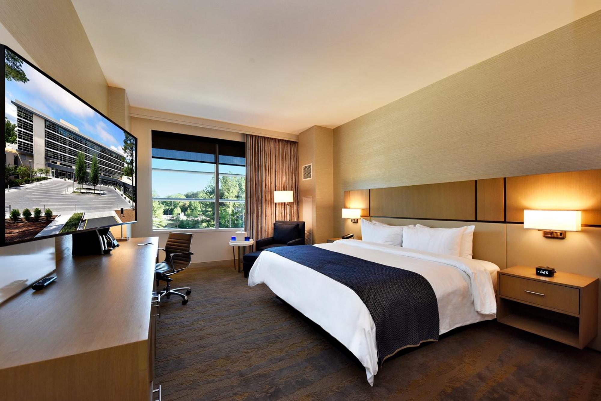 A guest bedroom with a large bed, light wood paneling, wall lamps, and a wide window overlooking trees.