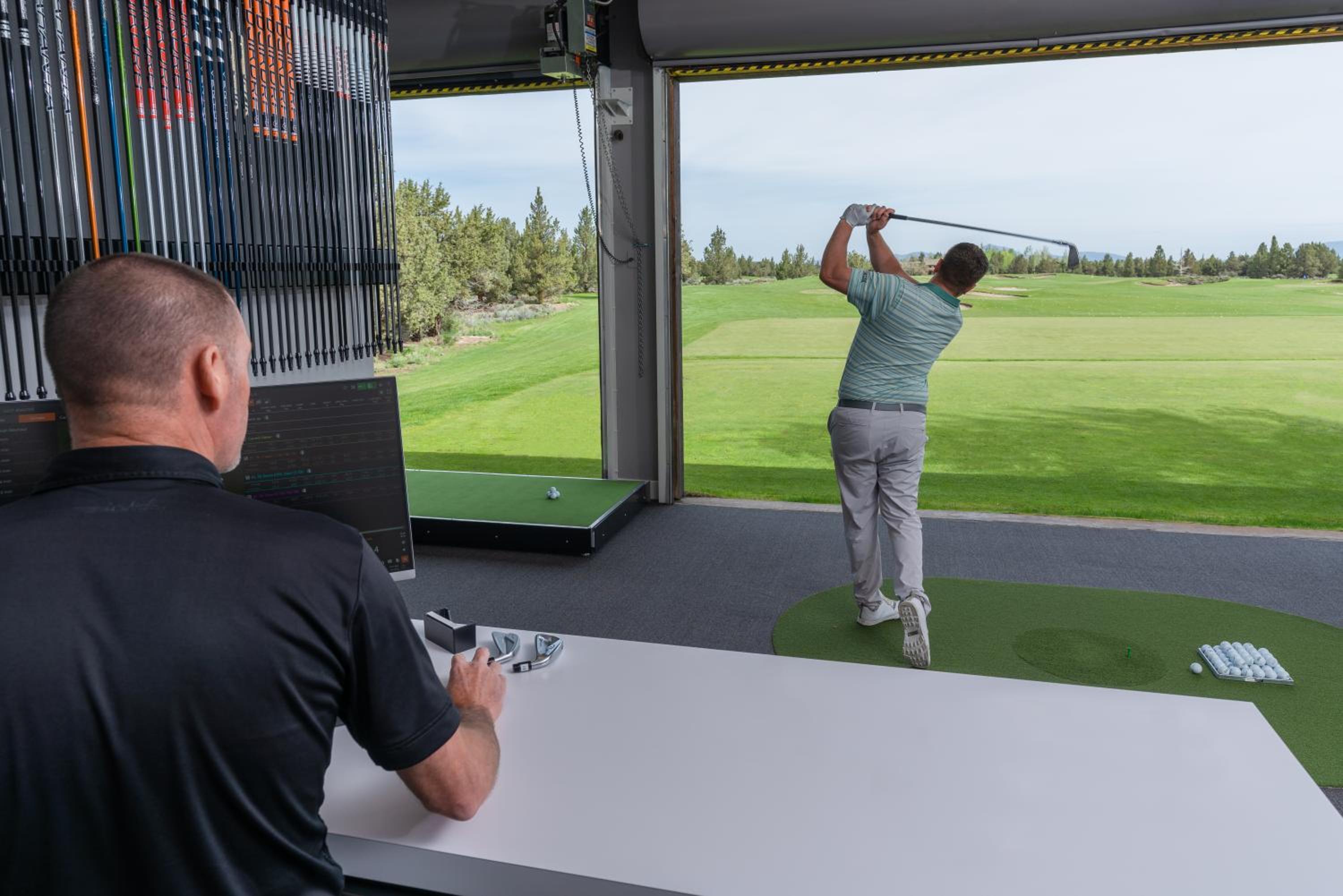 A golfer swings toward an open driving range while an instructor watches from a desk beside club shafts.