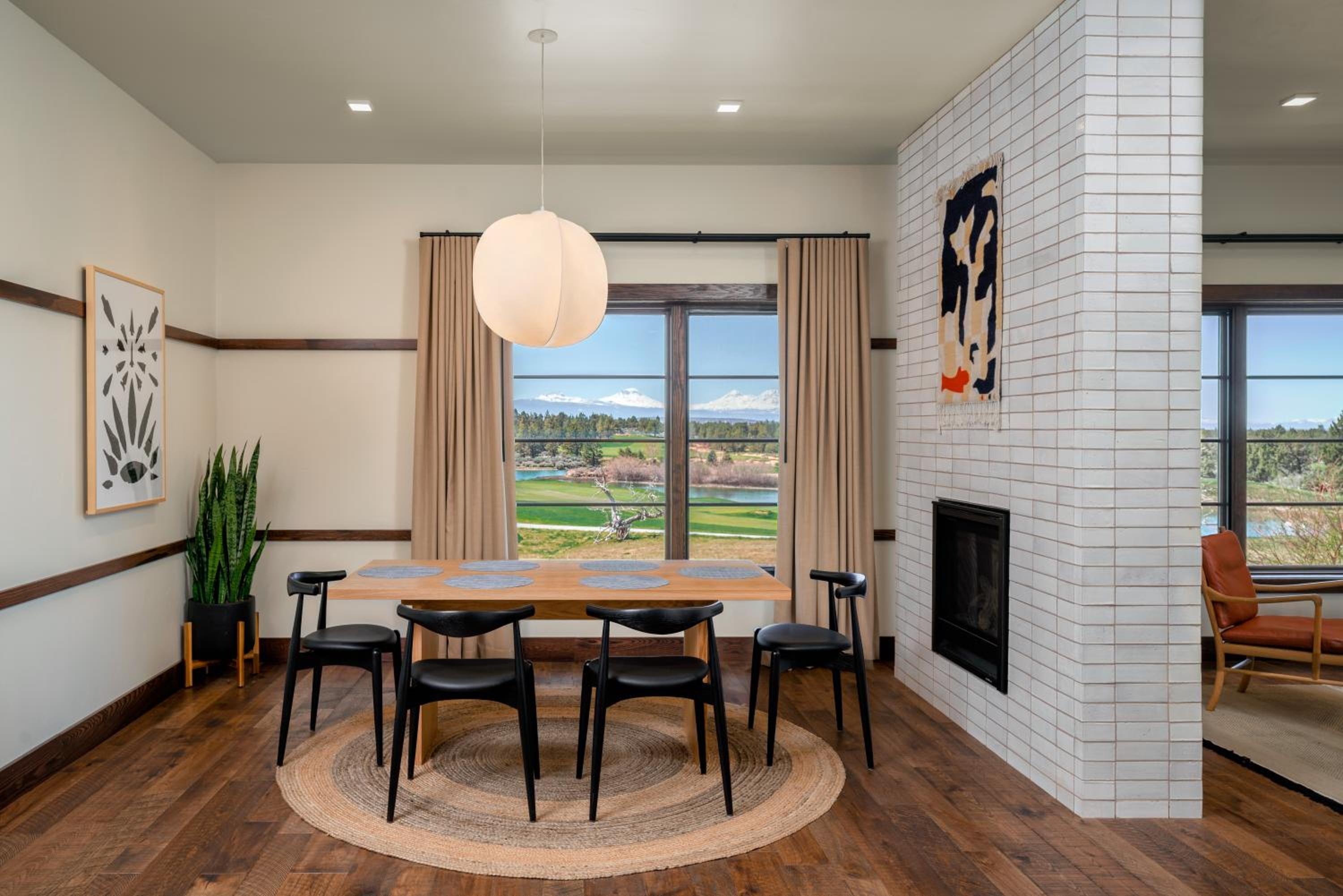 Dining area with a wooden table, round pendant light, white-tiled fireplace wall, and window view.