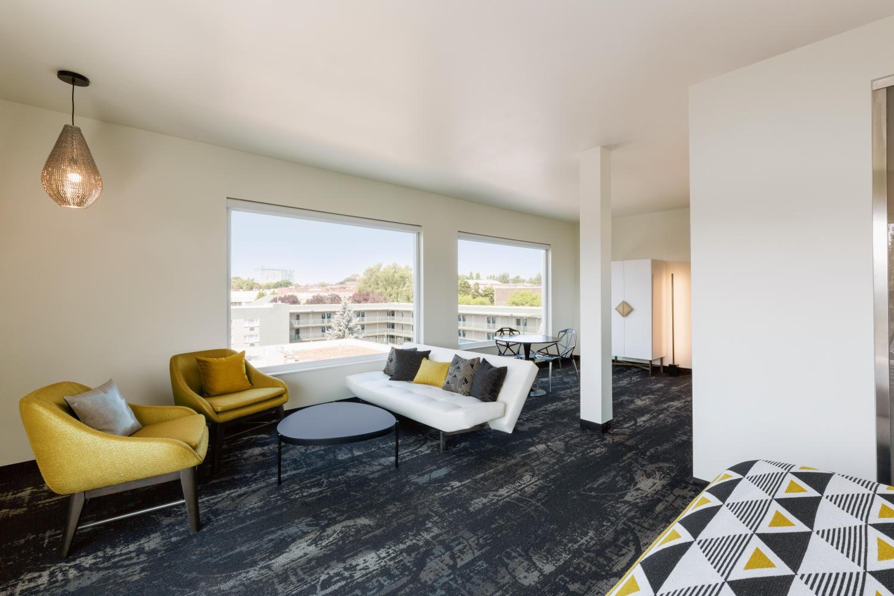 A bright sitting area with large picture windows, a white sofa, mustard armchairs, and patterned dark carpet.