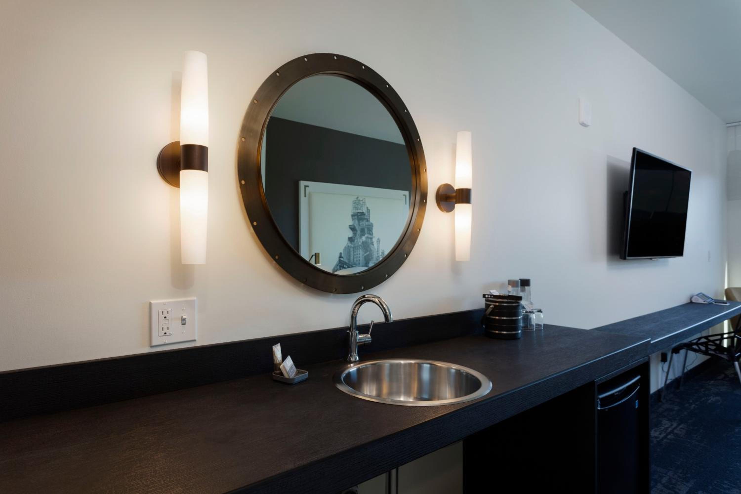 A dark wet bar counter with a round metal porthole mirror, stainless steel sink, and glowing wall sconces.