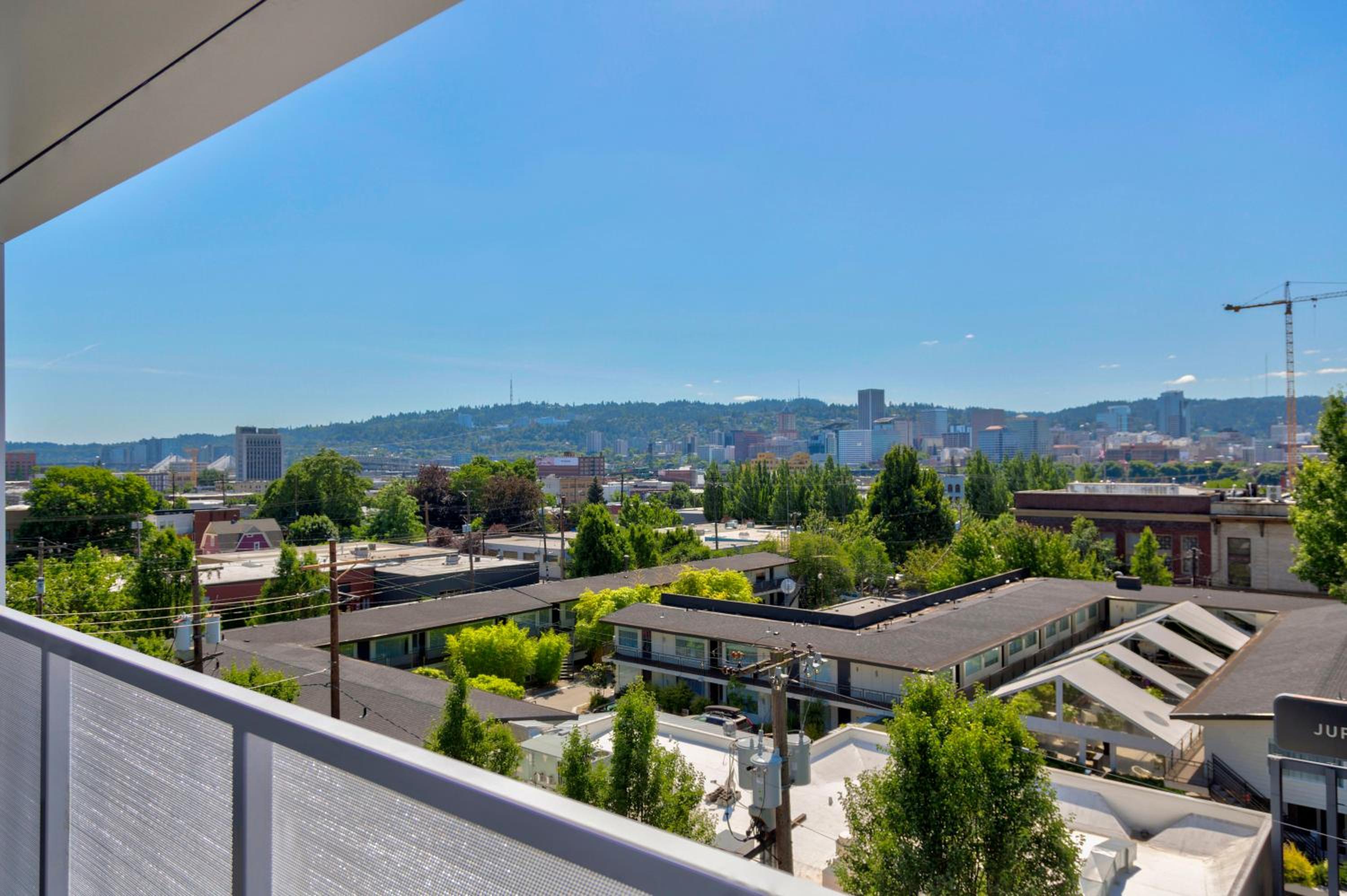 Balcony view looking over low-rise rooftops, green treetops, and a distant city skyline under a clear sky.