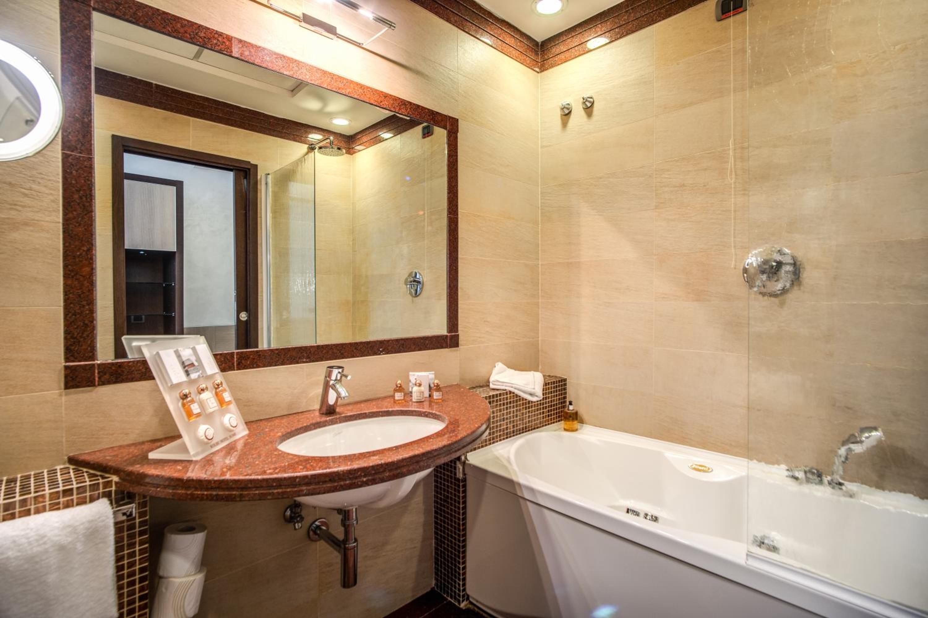 Hotel bathroom with a reddish-brown stone vanity, large mirror, and white bathtub set against beige tiled walls.