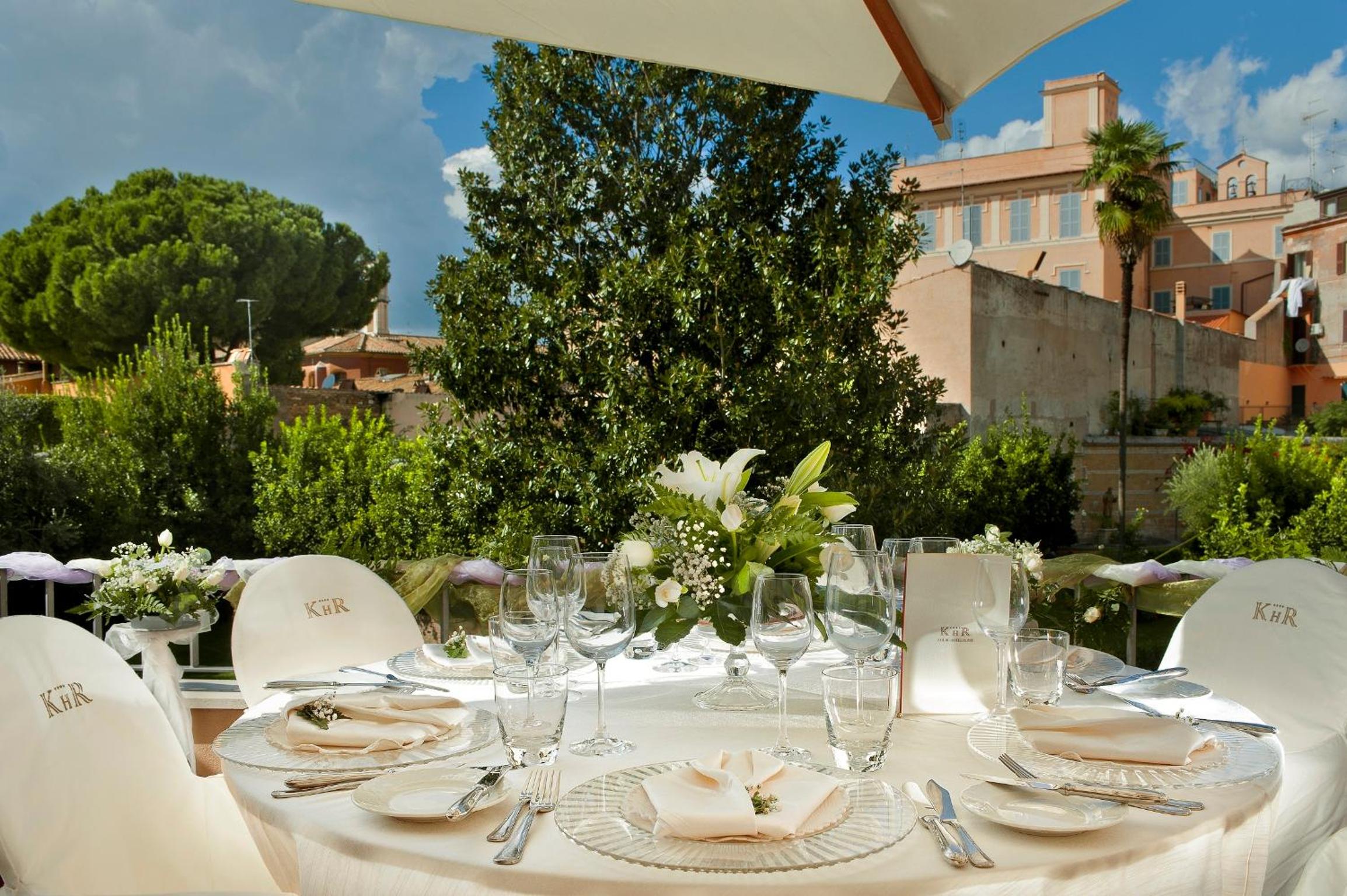 Round dining table laid with white linens, glassware, and floral centerpiece on an outdoor terrace framed by trees.