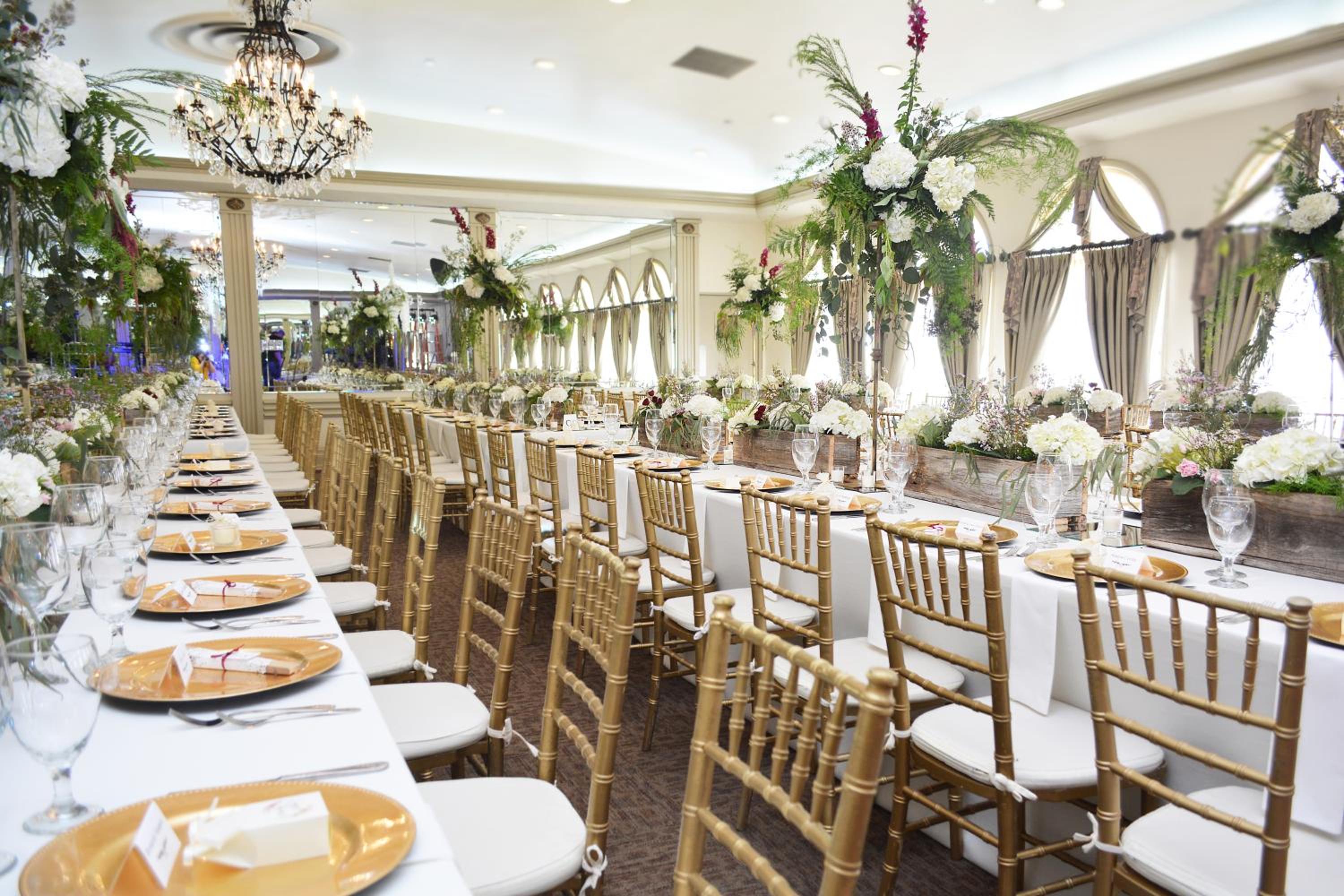 An indoor event room arranged with banquet tables, tall floral centerpieces, gold chairs, and a chandelier.