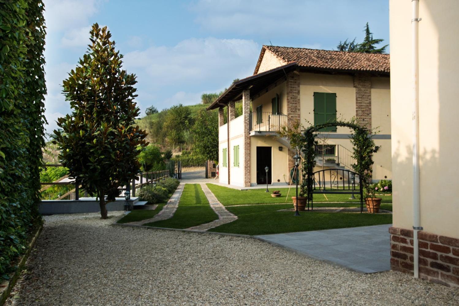 A lawn and gravel driveway leading toward a country house with green shutters, brick accents, and a tiled roof.