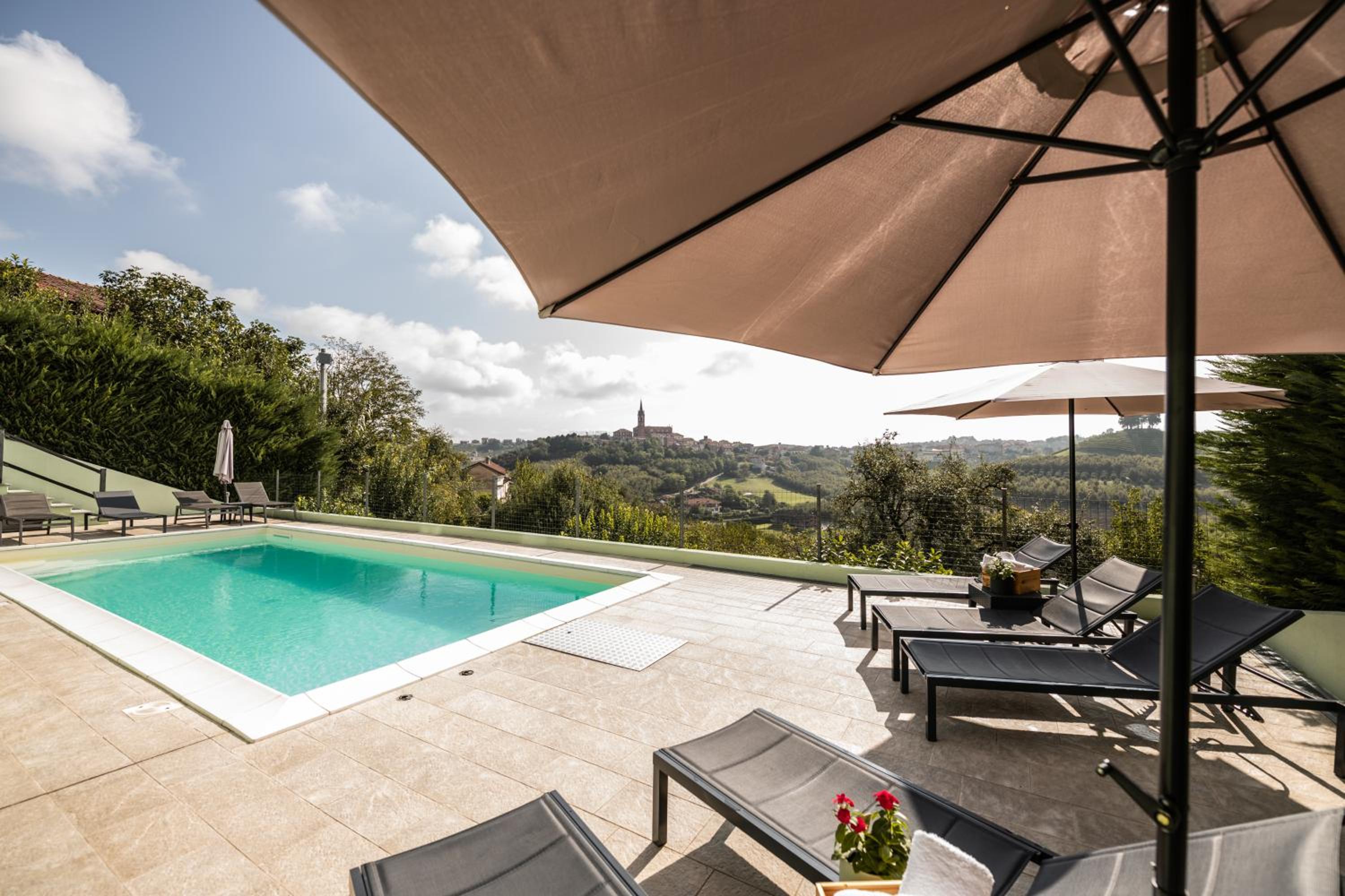 A swimming pool and sun loungers on a paved terrace overlooking rolling vineyard hills and a village.